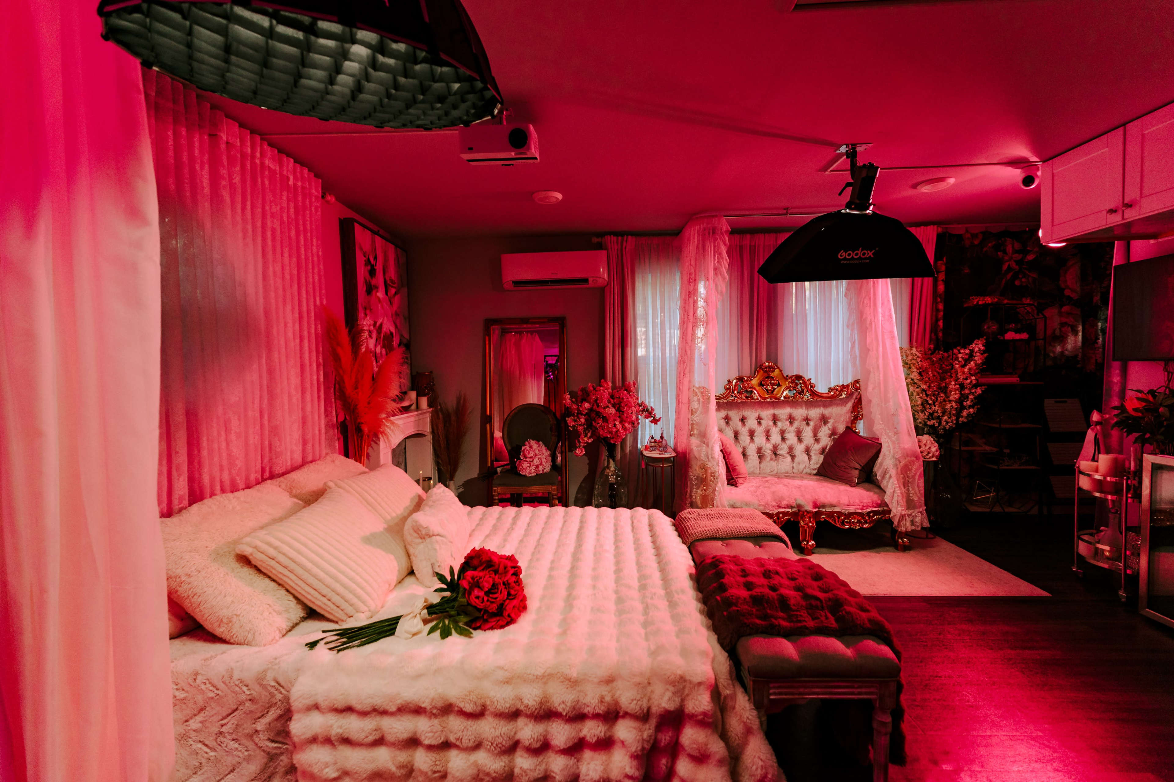 The image shows a stylish bedroom decorated with pink lighting, featuring a large bed covered with a plush blanket and a vintage-style sofa.
