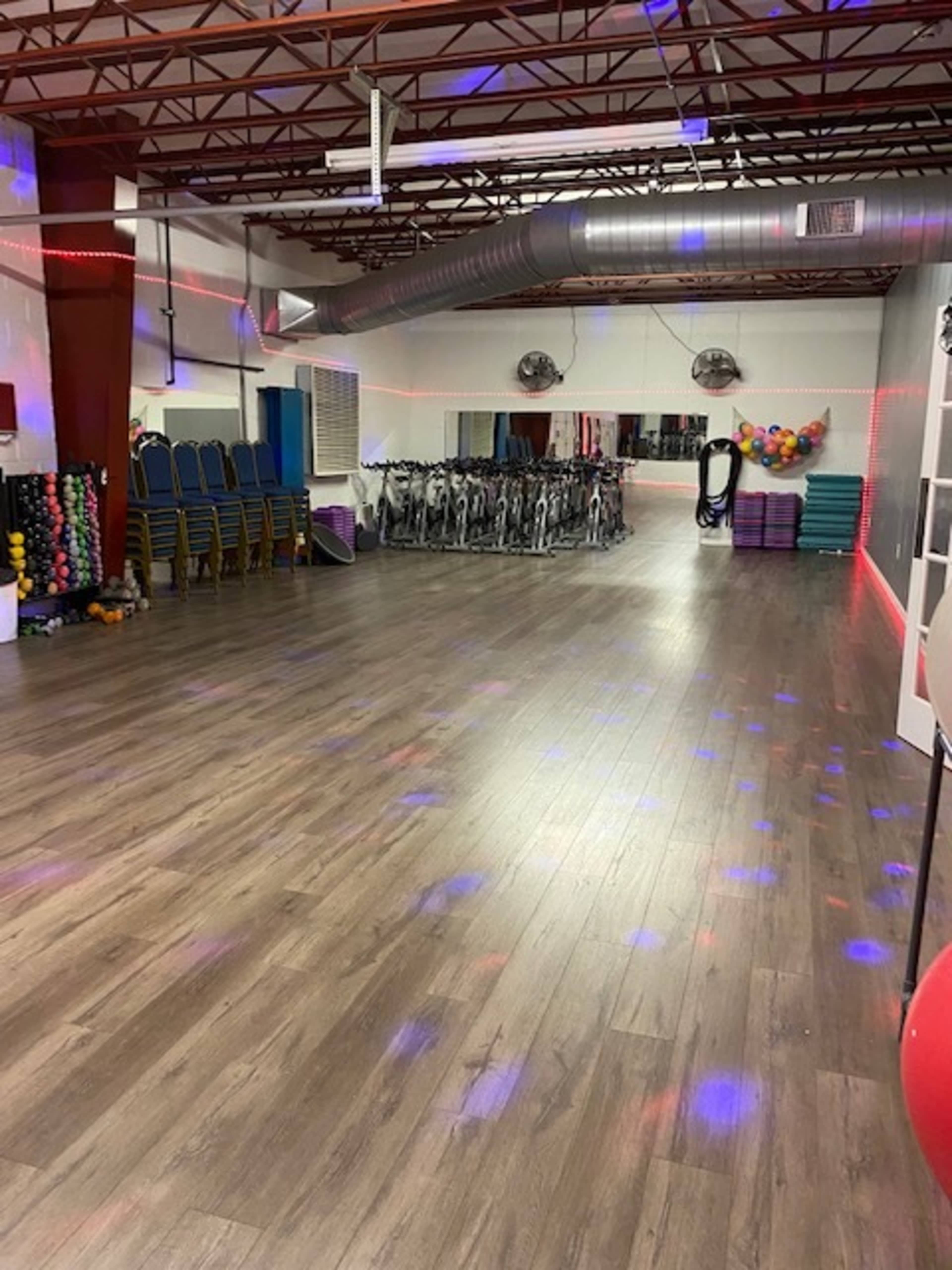 A fitness studio with exercise equipment, including stationary bikes, stacked exercise mats, and weights, under colored lights on a wooden floor.