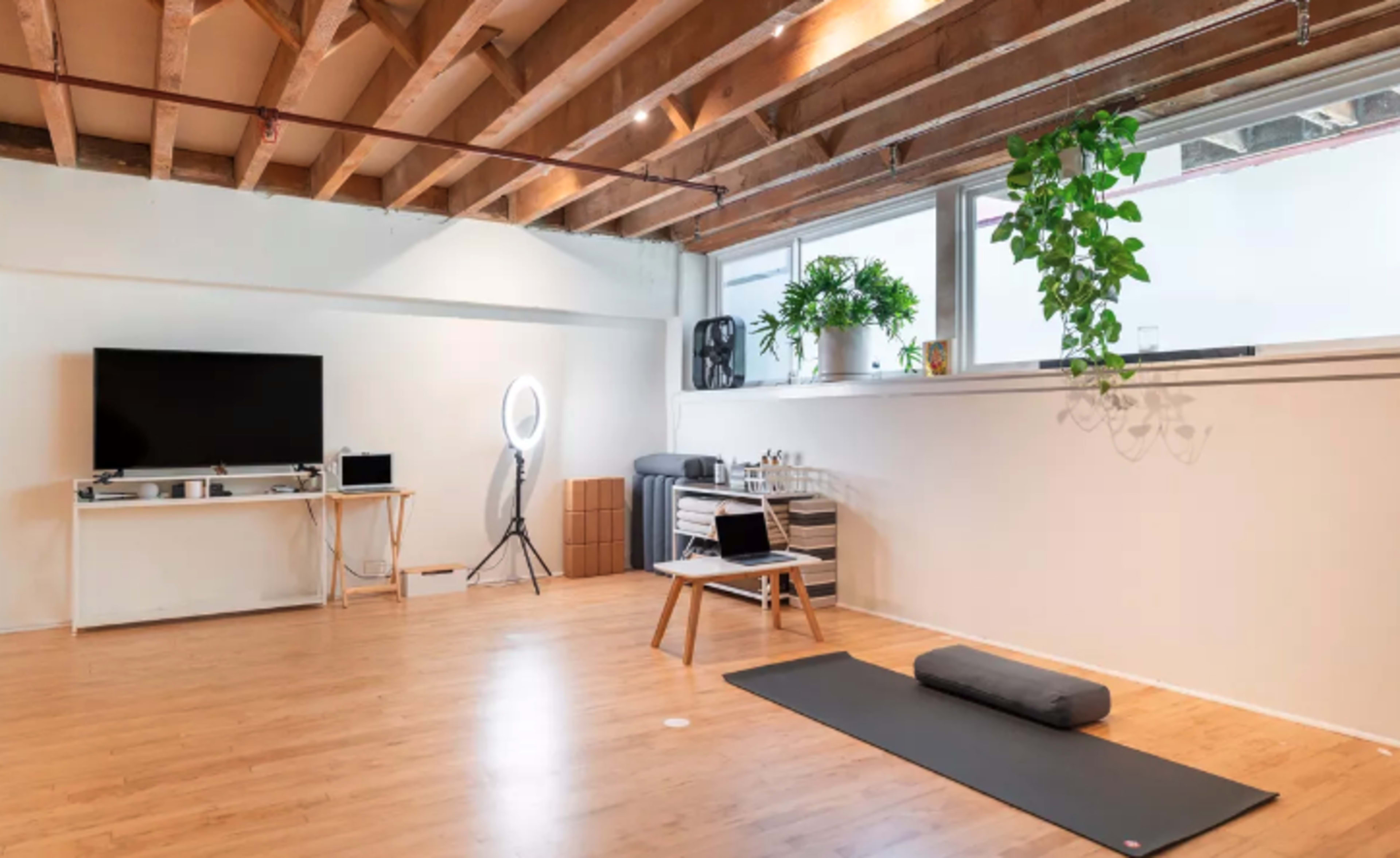 A spacious room with wooden beams, a television on a white stand, a ring light, and a yoga mat laid out on the floor.