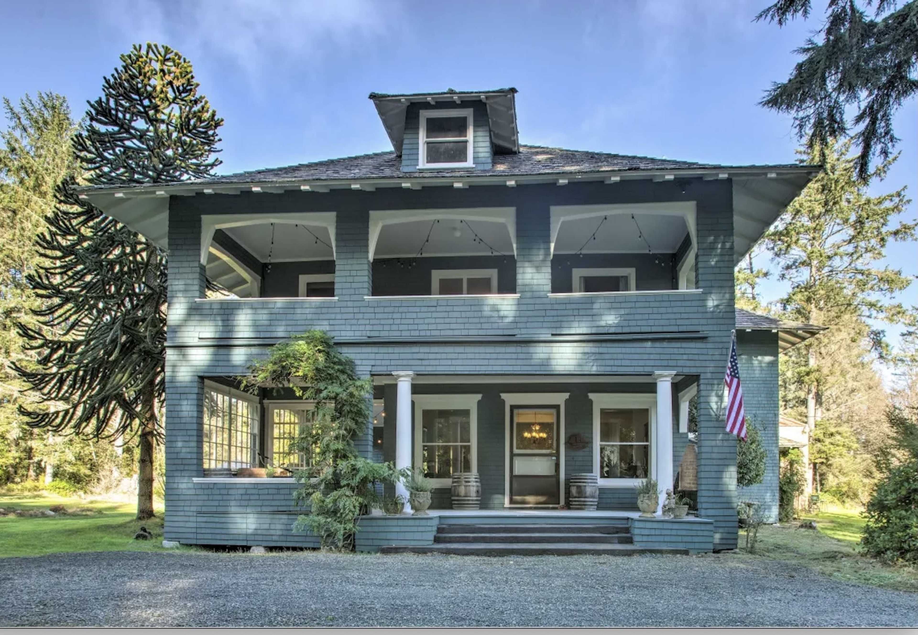 A large, two-story, blue-painted house with a covered porch, multiple windows, and an American flag hanging at the entrance.