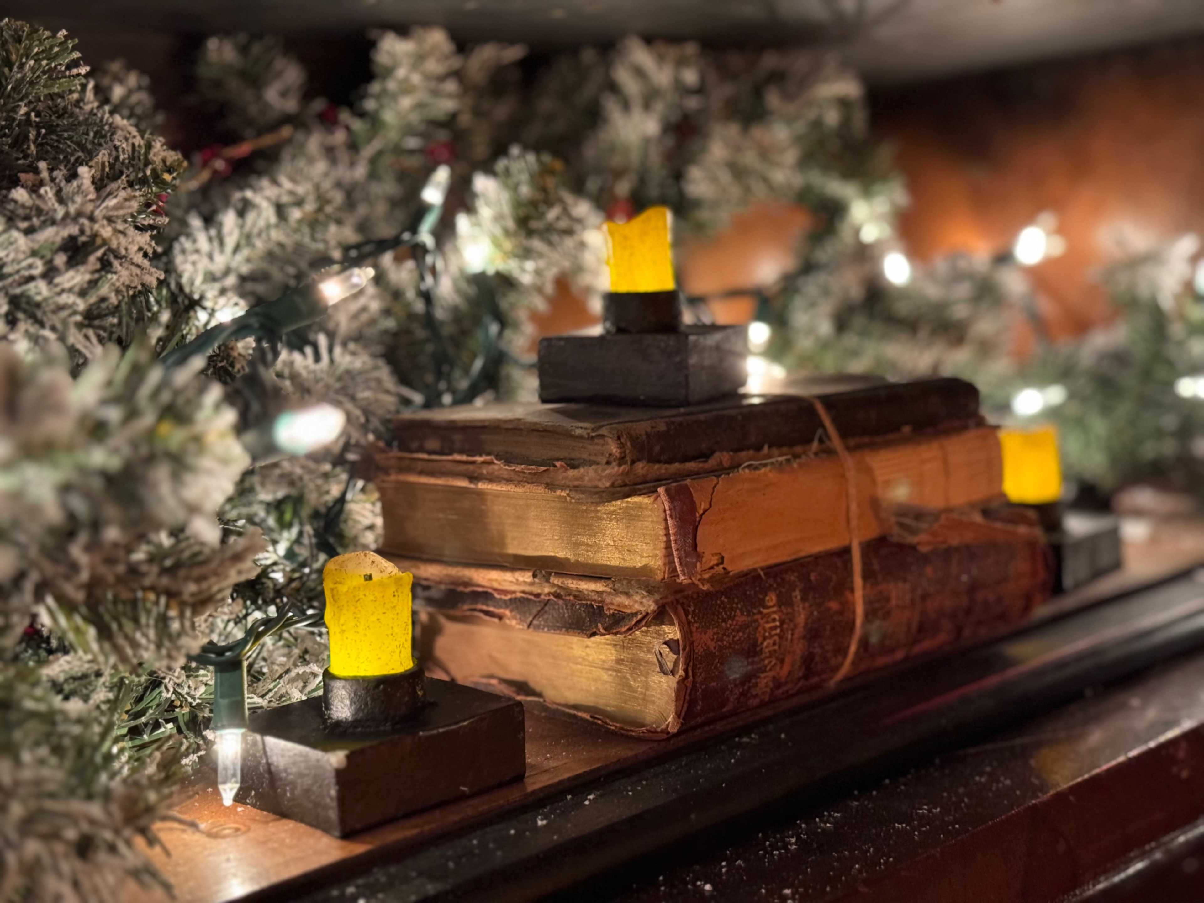 A stack of old books tied with rope rests on a shelf adorned with garlands and small yellow candle lights.