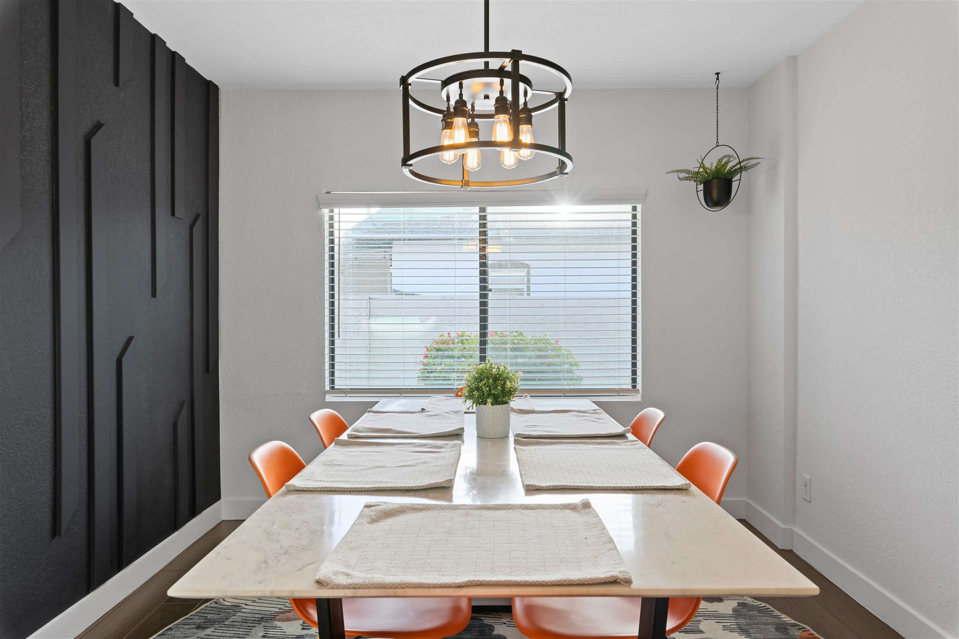 A modern dining area features a long table with orange chairs, a pendant light, and a window with blinds allowing natural light to enter.