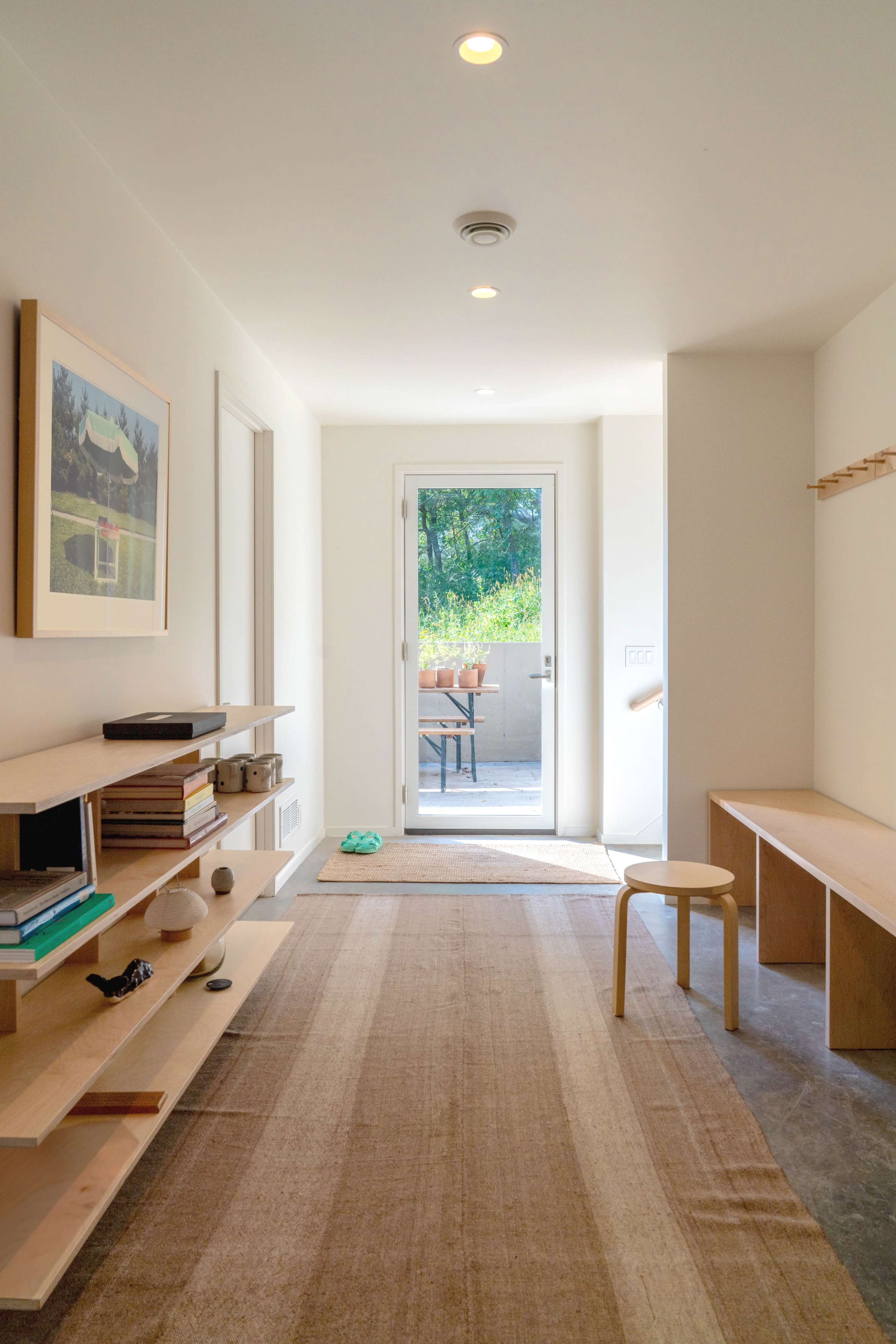 The image shows a modern interior hallway leading to a patio, featuring a minimalist design with a wooden shelf, a stool, and a large door that opens to a green outdoor space.