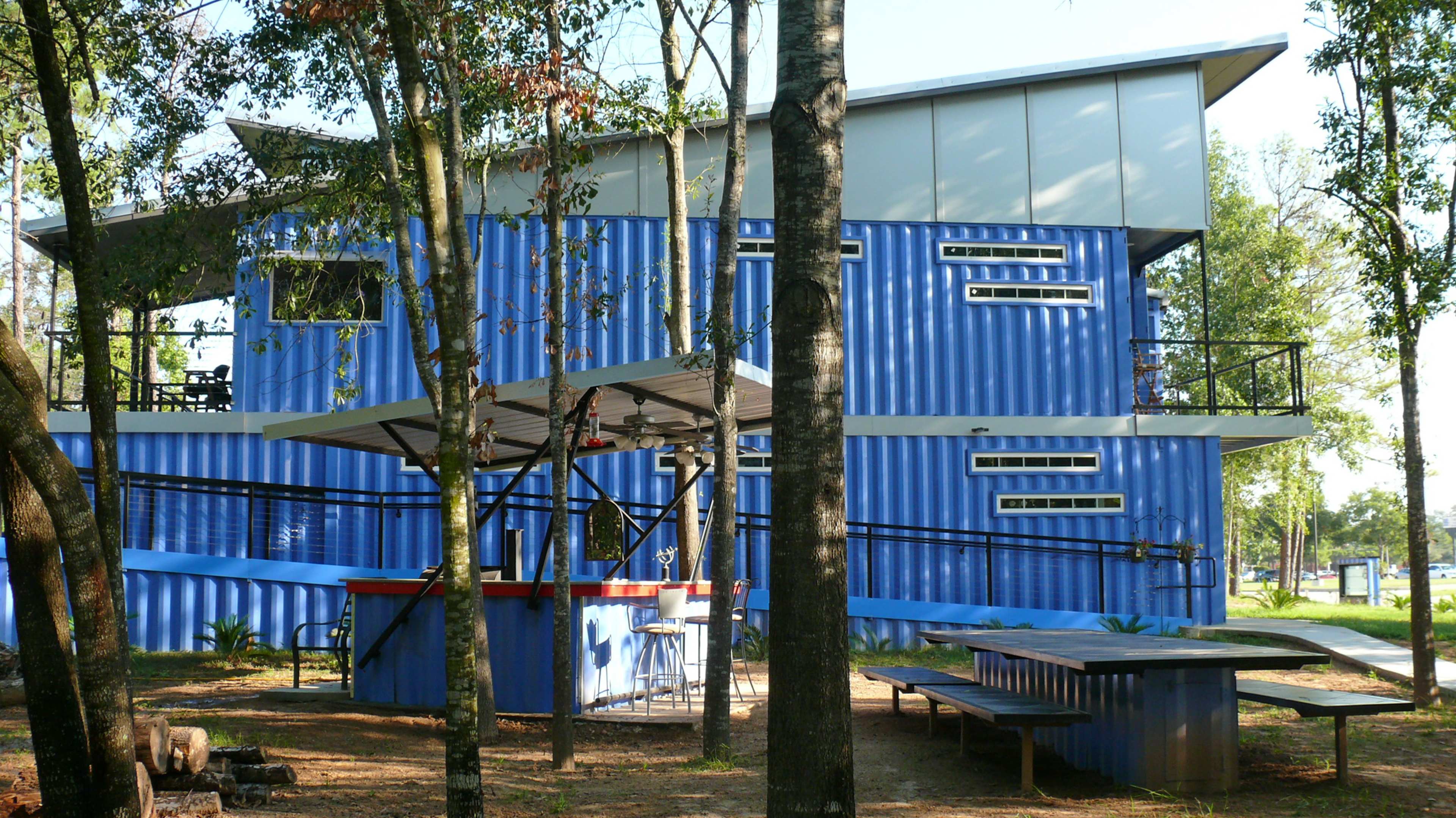 A modern blue corrugated metal house surrounded by trees, with an outdoor bar and picnic area in the foreground.