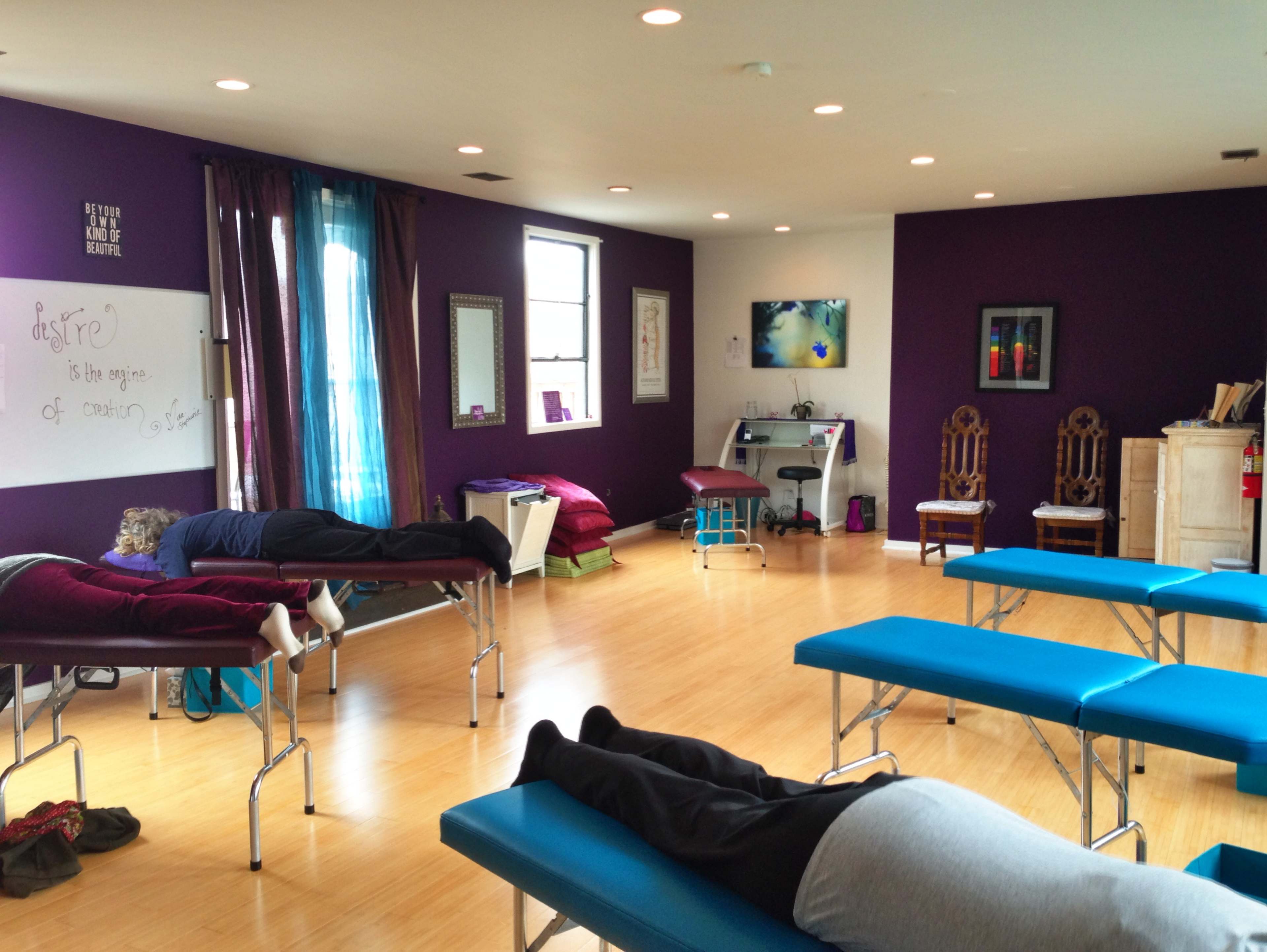A brightly lit therapy room with several treatment tables occupied by individuals lying down, surrounded by purple walls and calming decor.