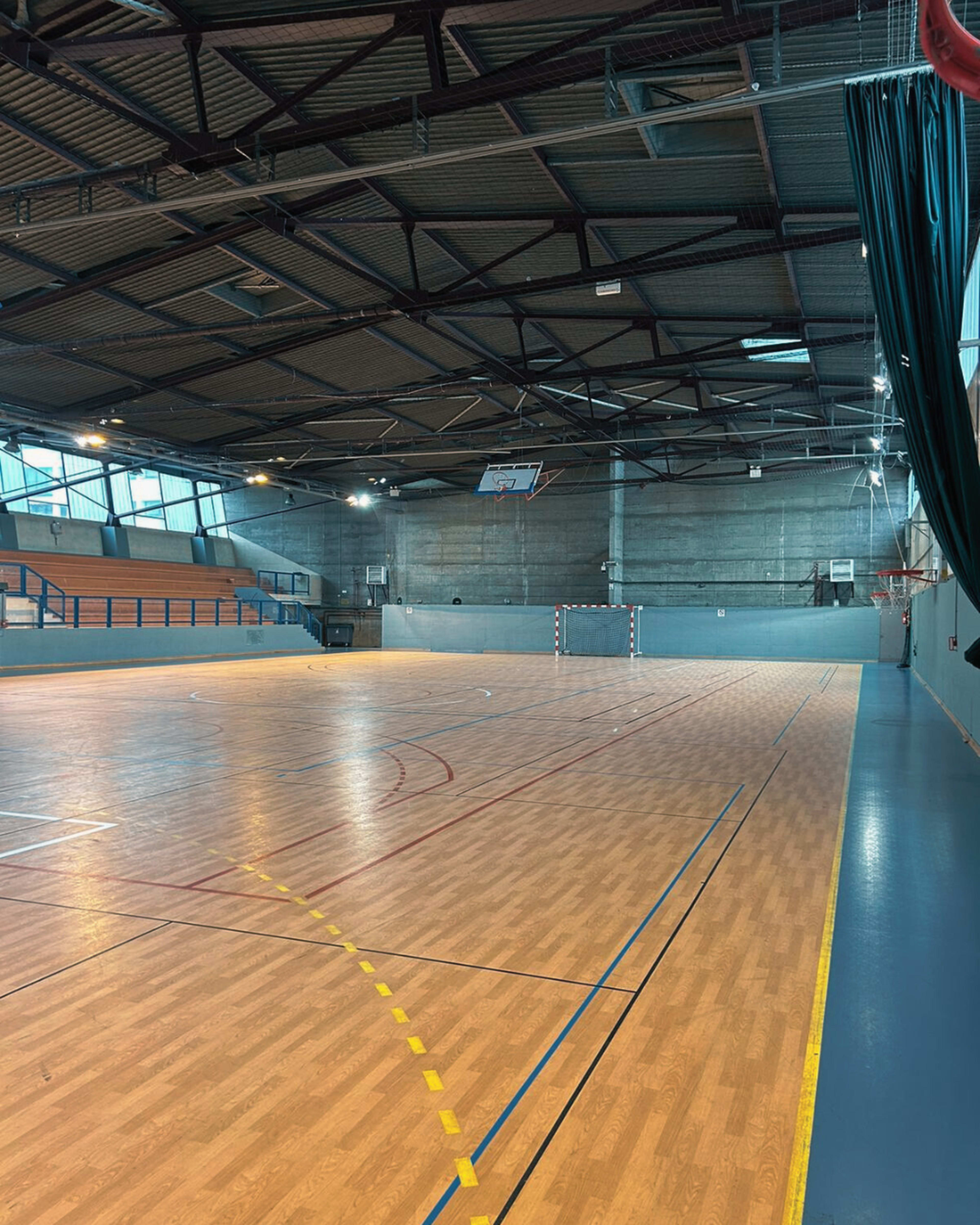 A spacious indoor sports hall with a wooden floor, basketball hoop, and goal posts.