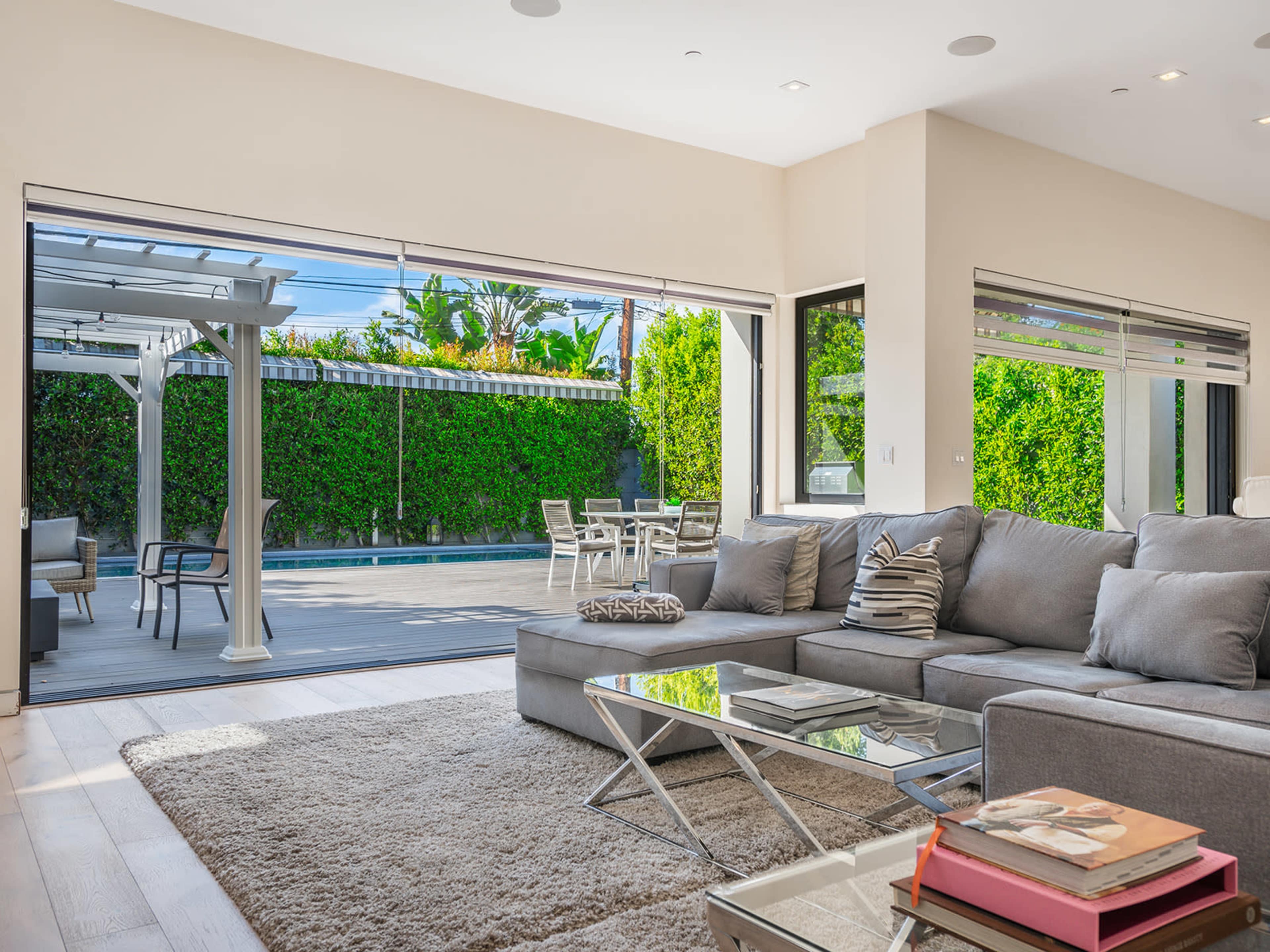 A modern living room with a gray sectional sofa overlooks a patio and pool surrounded by greenery through large sliding glass doors.