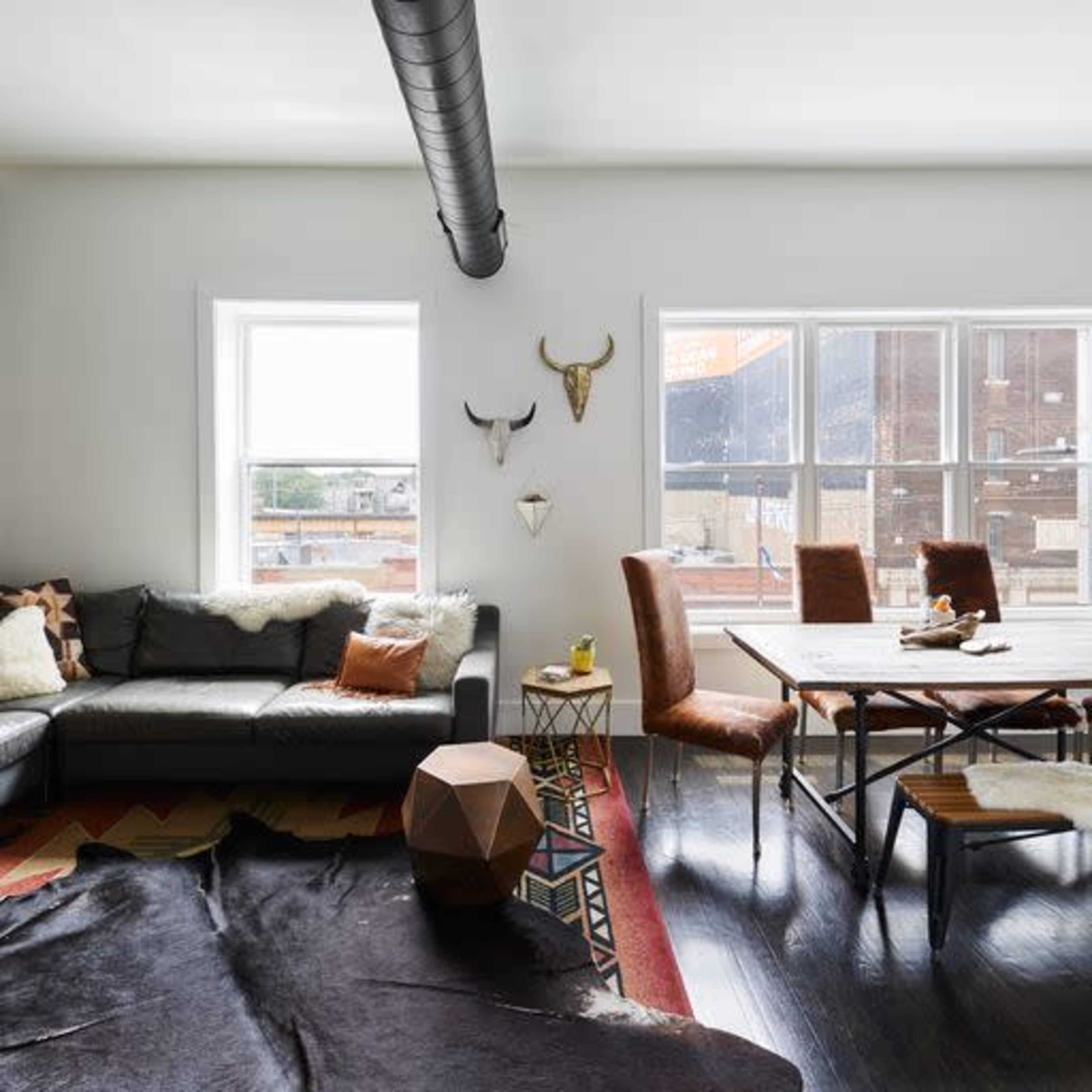 A modern living room features a leather sofa, wooden dining table, and decorative accents, with large windows allowing natural light to illuminate the space.