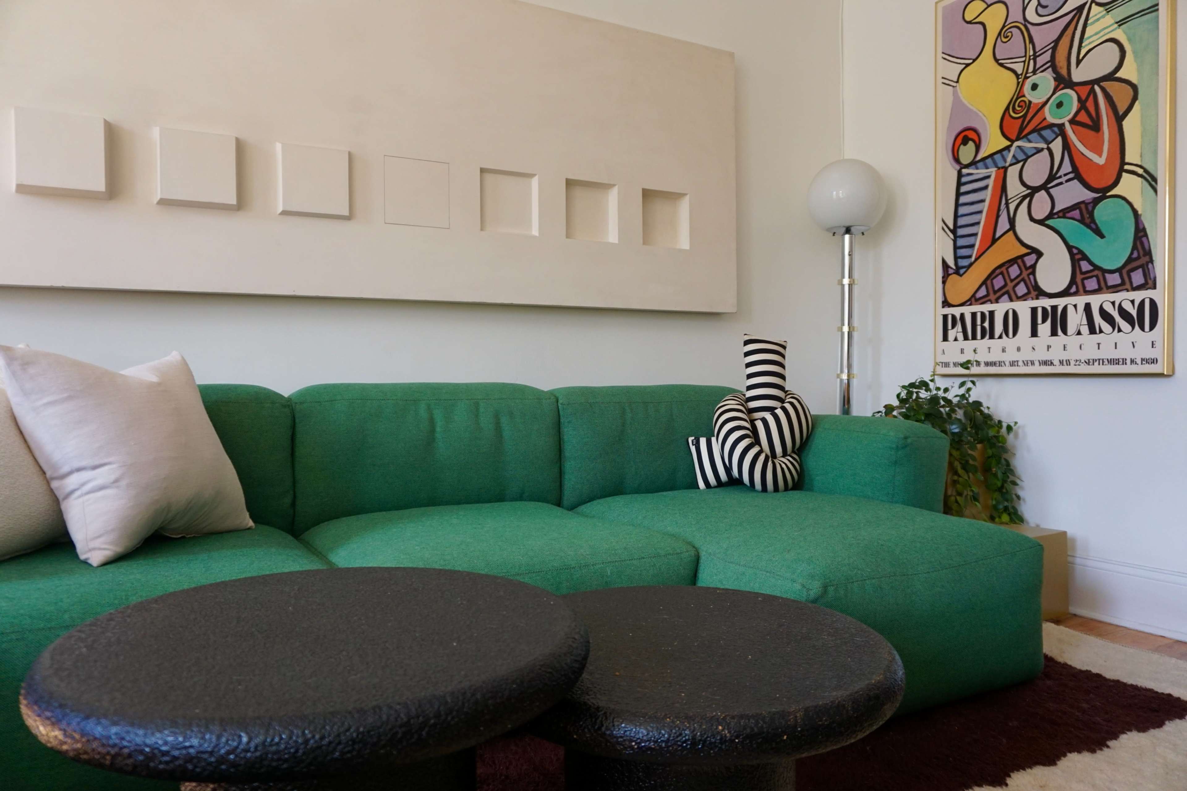 A green sectional sofa is positioned in a room with a patterned artwork and two black round tables in front.