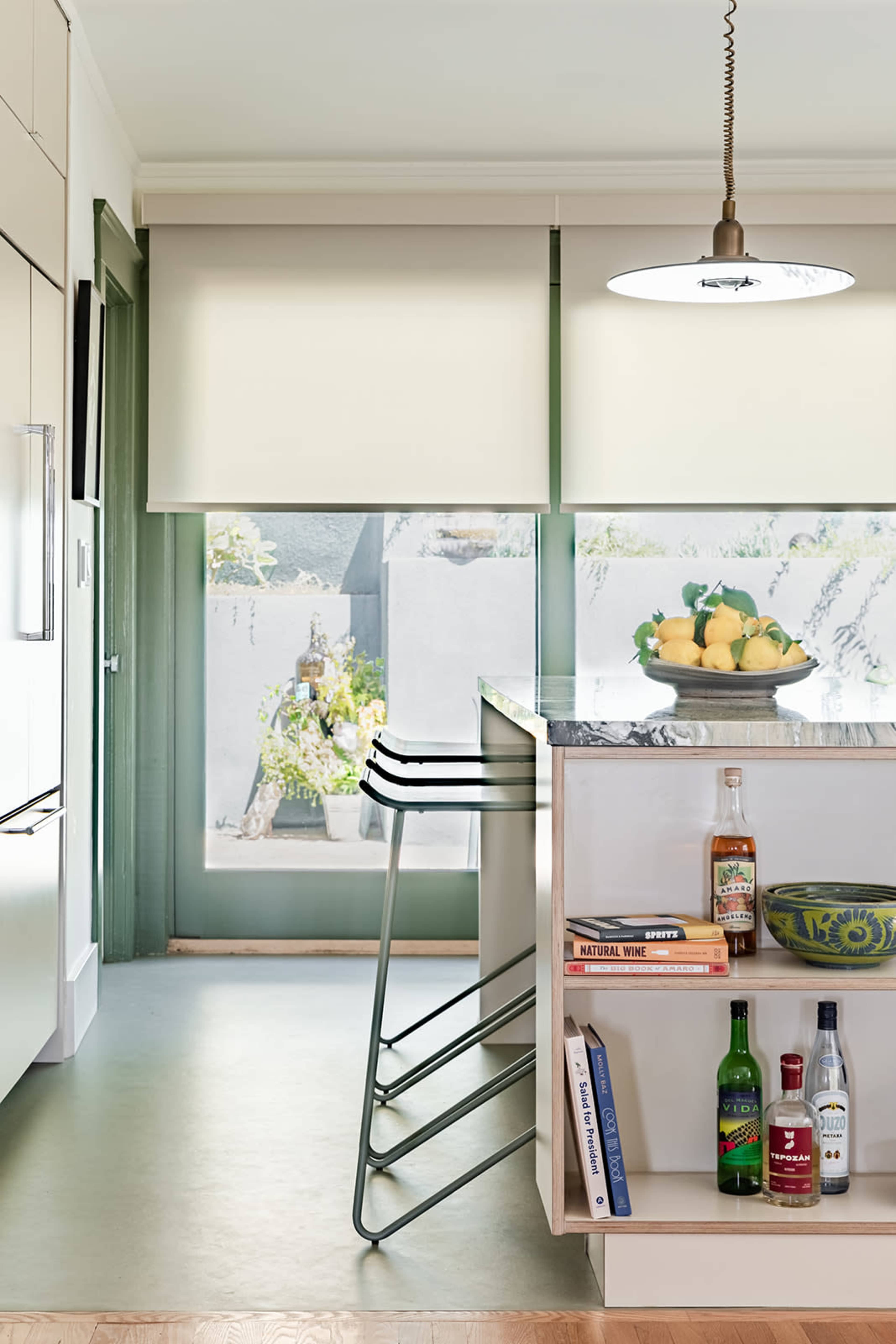 The kitchen features a modern bar countertop with metal stools, a fruit bowl, and a view of a landscaped outdoor area through large windows.
