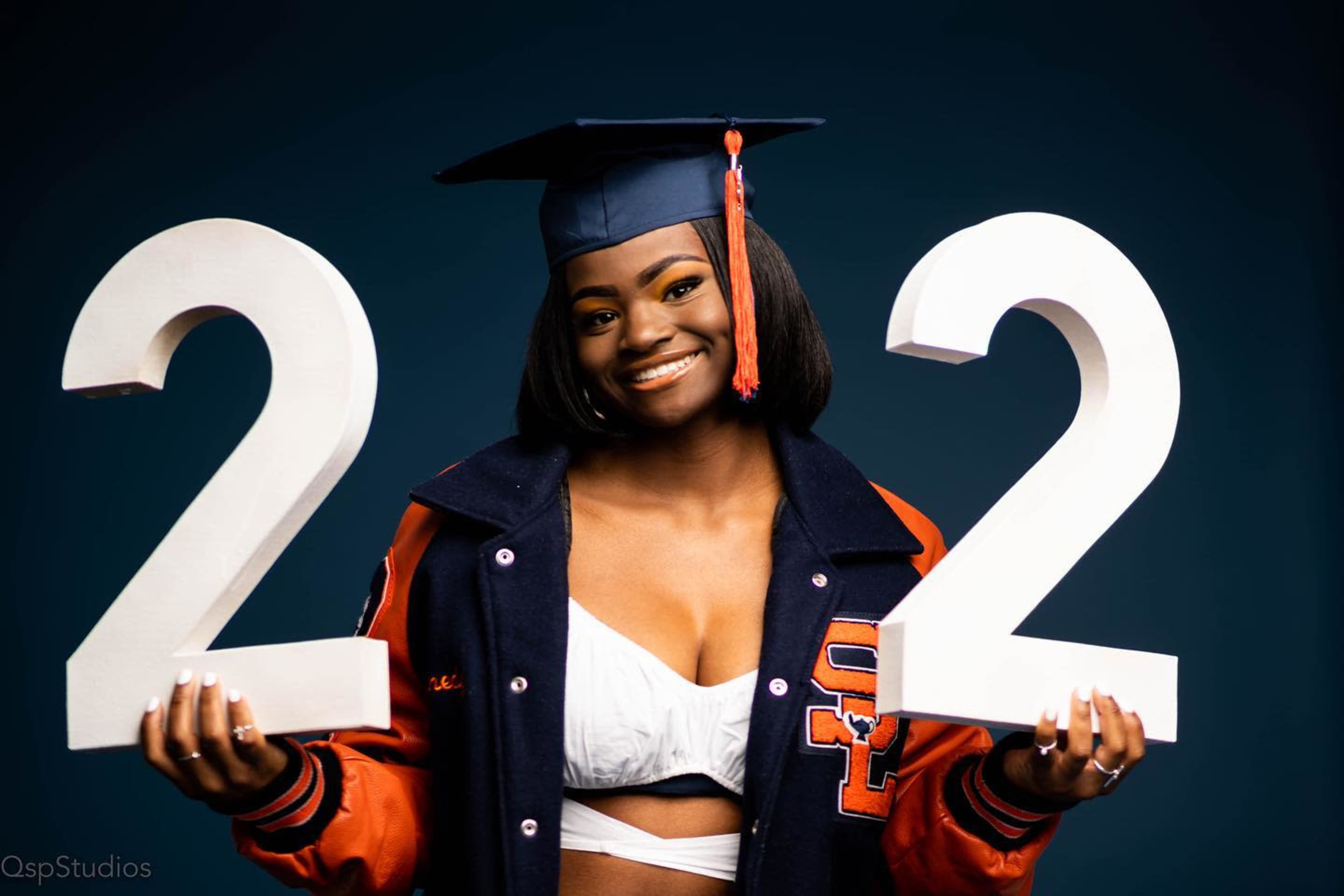 A graduate wearing a cap and gown holds up two large foam numbers "2" and "2."