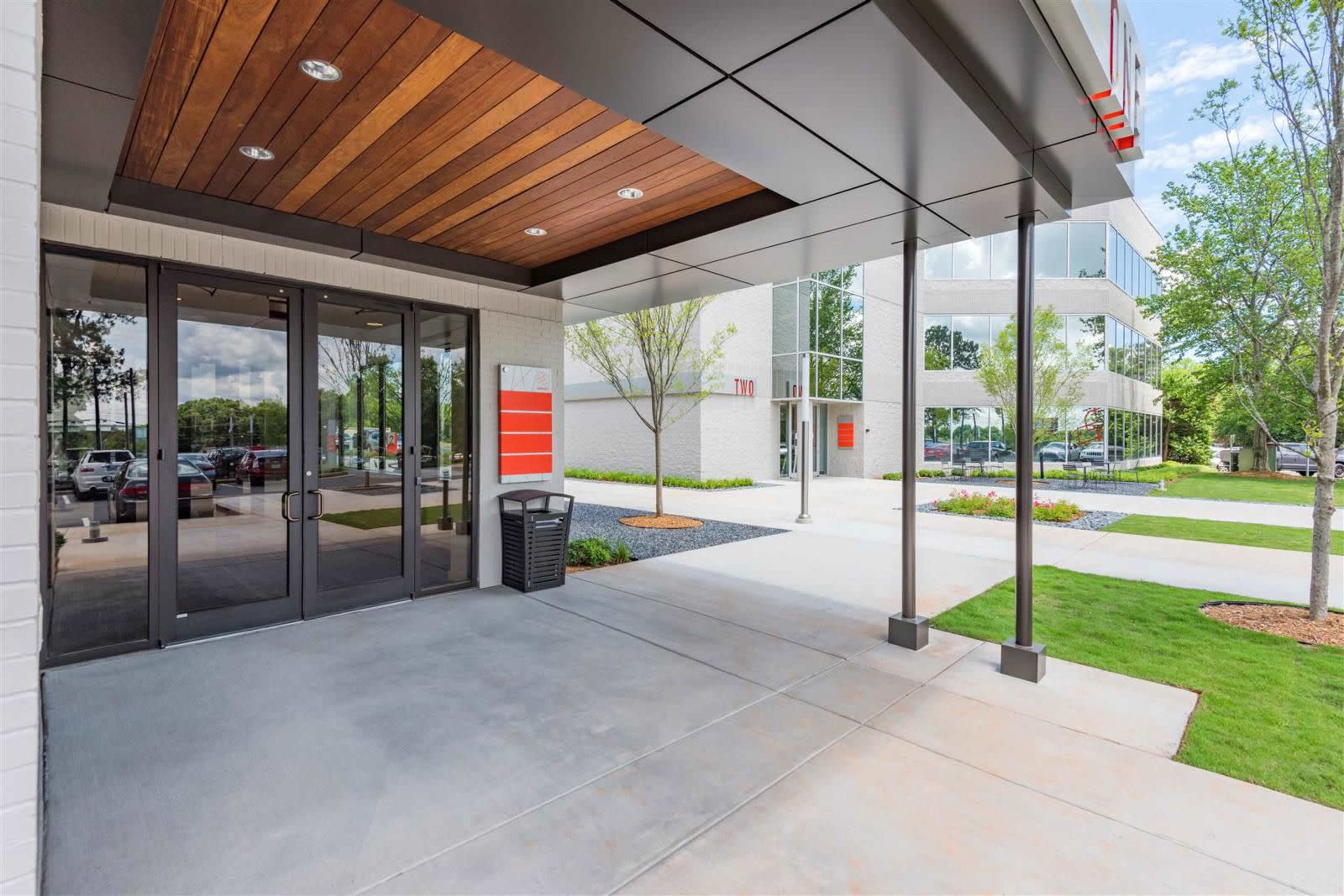 The image shows the entrance of a modern building with large glass doors, a concrete walkway, and landscaped areas with trees and shrubs.