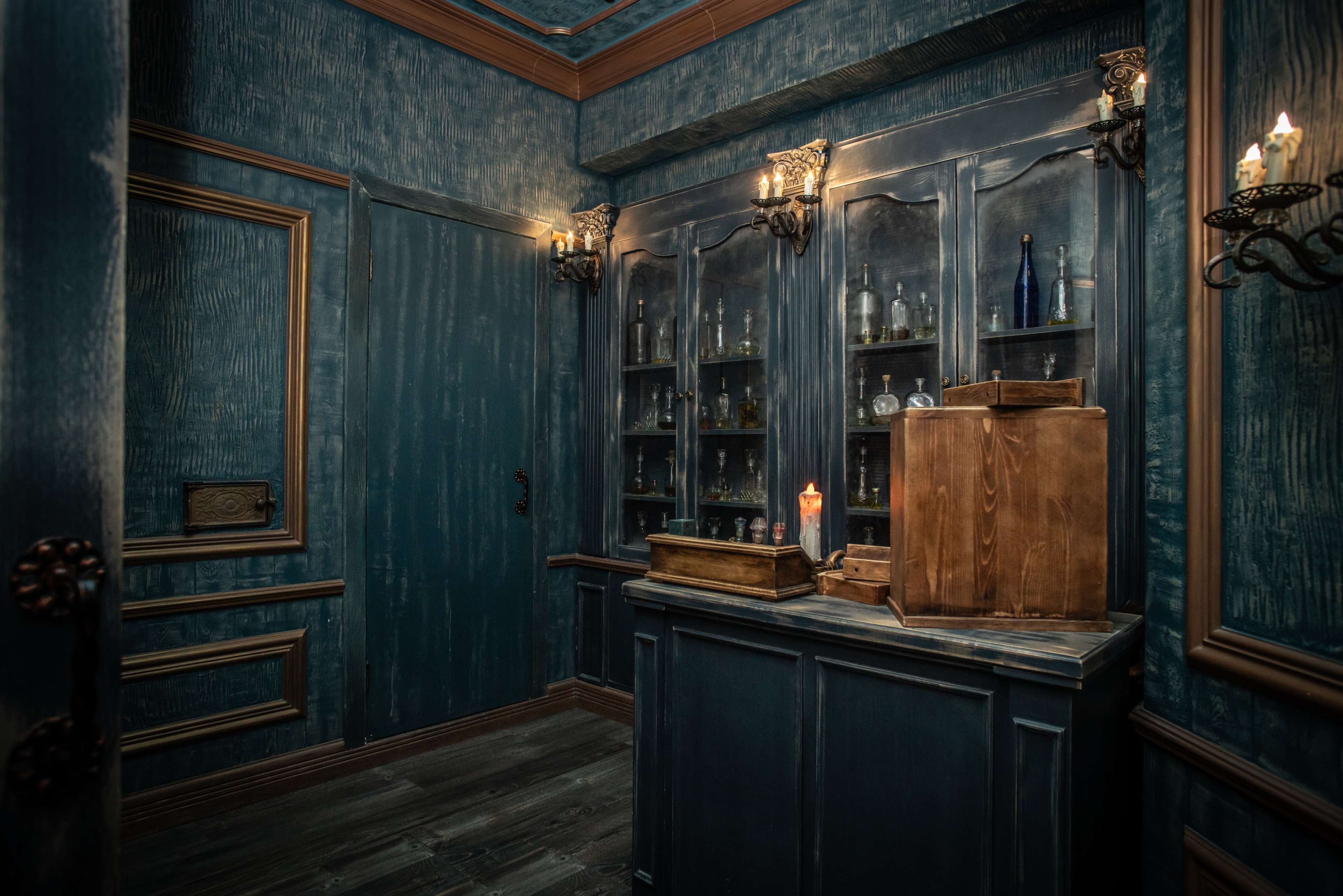 A dimly lit, vintage-style room with dark blue walls, a wooden counter, and shelves filled with various glass bottles.