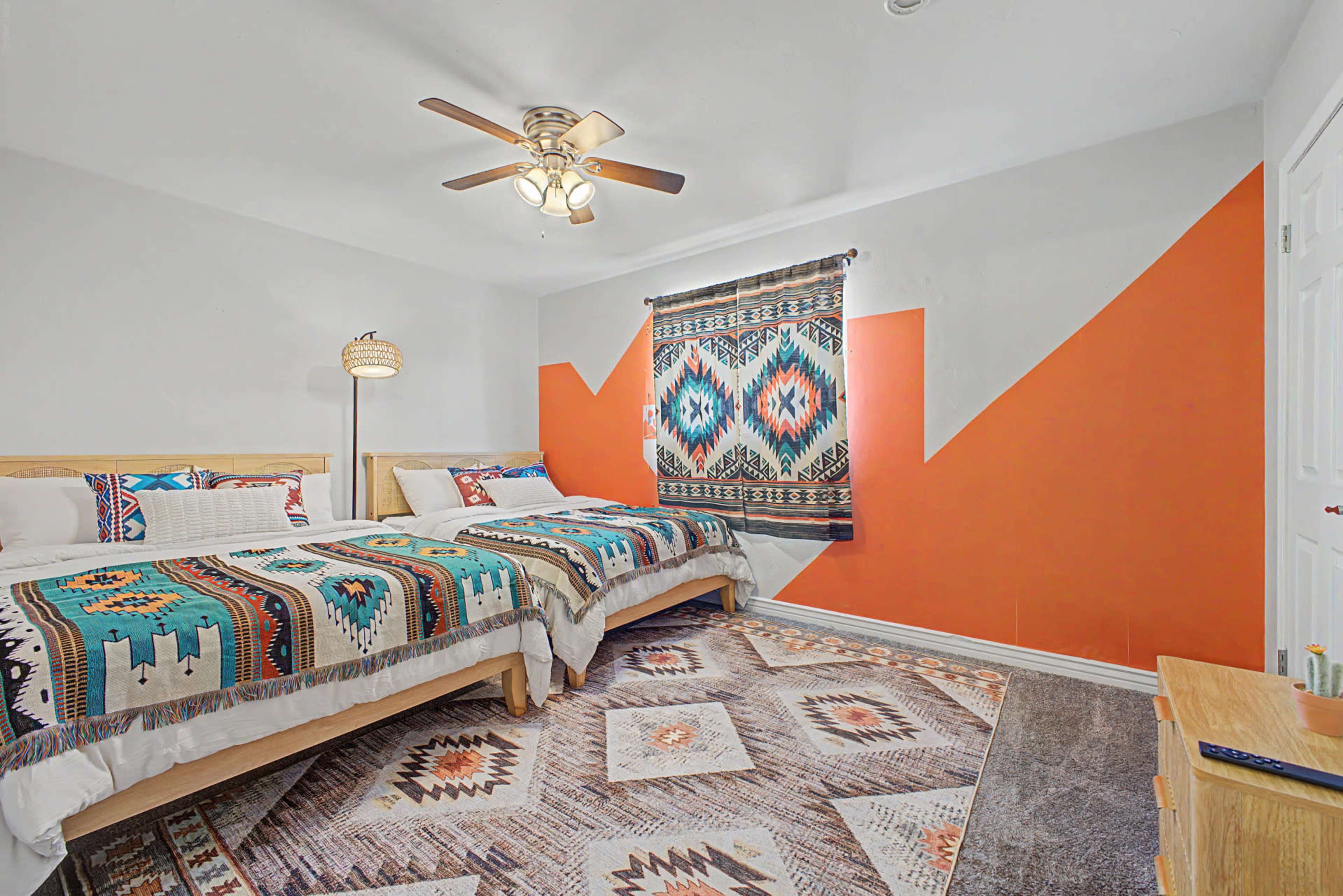 The image shows a brightly decorated bedroom featuring two twin beds with patterned blankets, a ceiling fan, and a colorful accent wall.