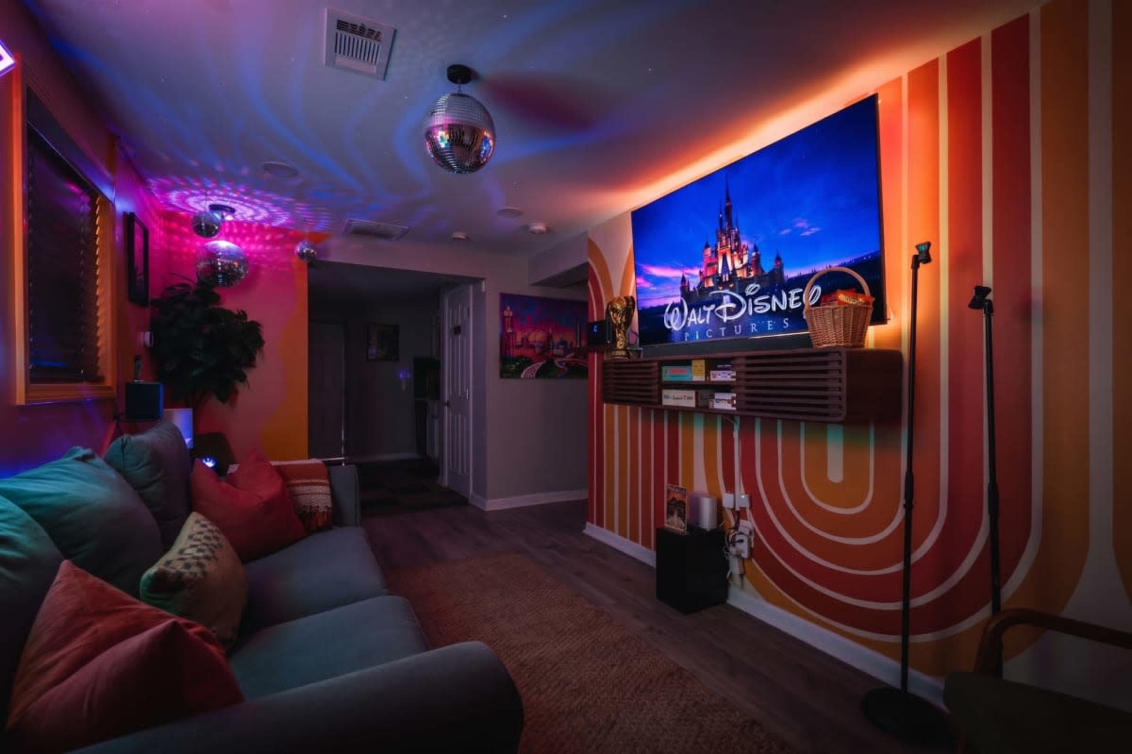 The image shows a colorful living room with a large television displaying a Disney logo, surrounded by vibrant wall decor and ambient lighting effects.