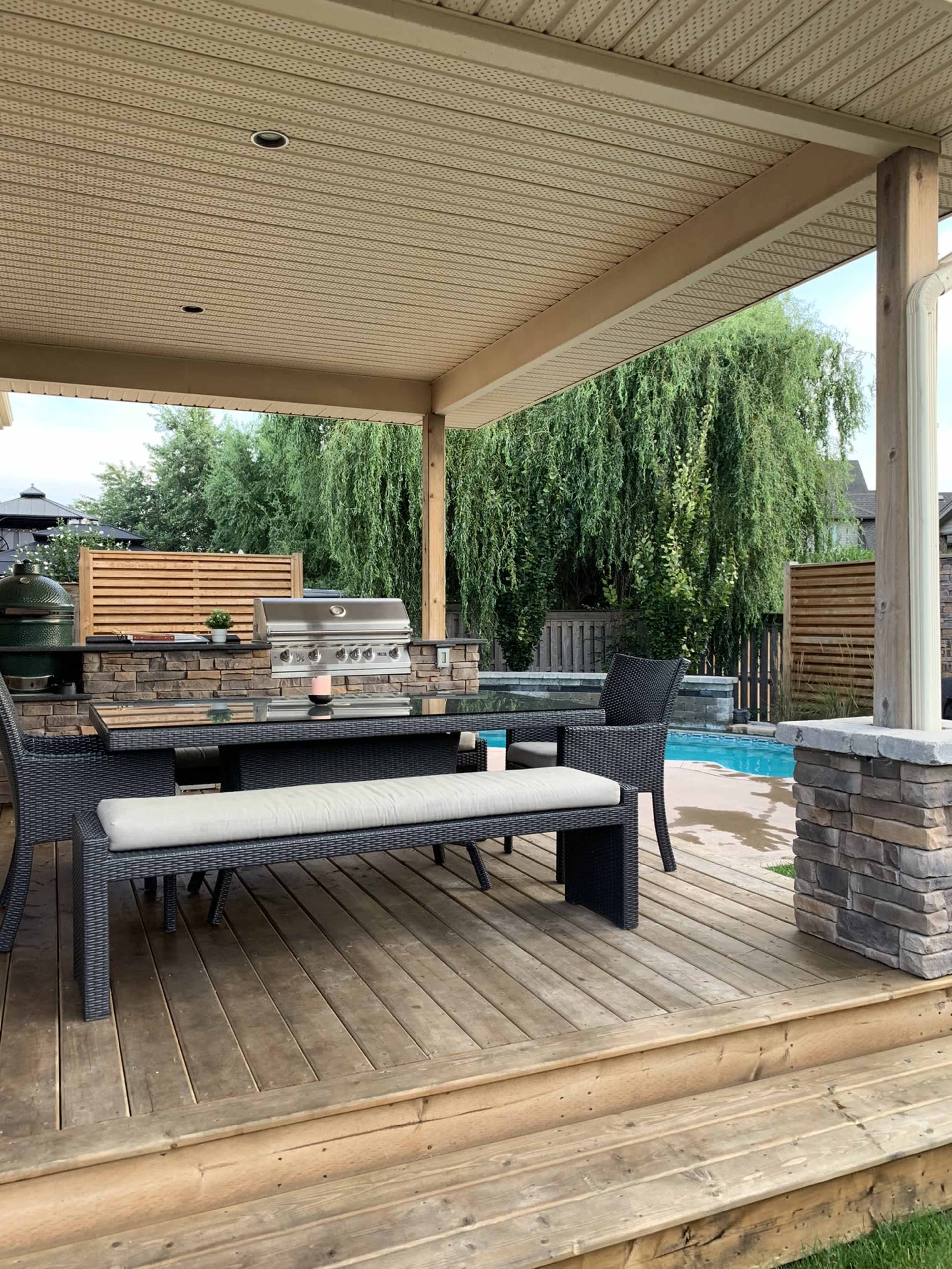 A patio area with a grill, a table with a bench, and a swimming pool in the background.