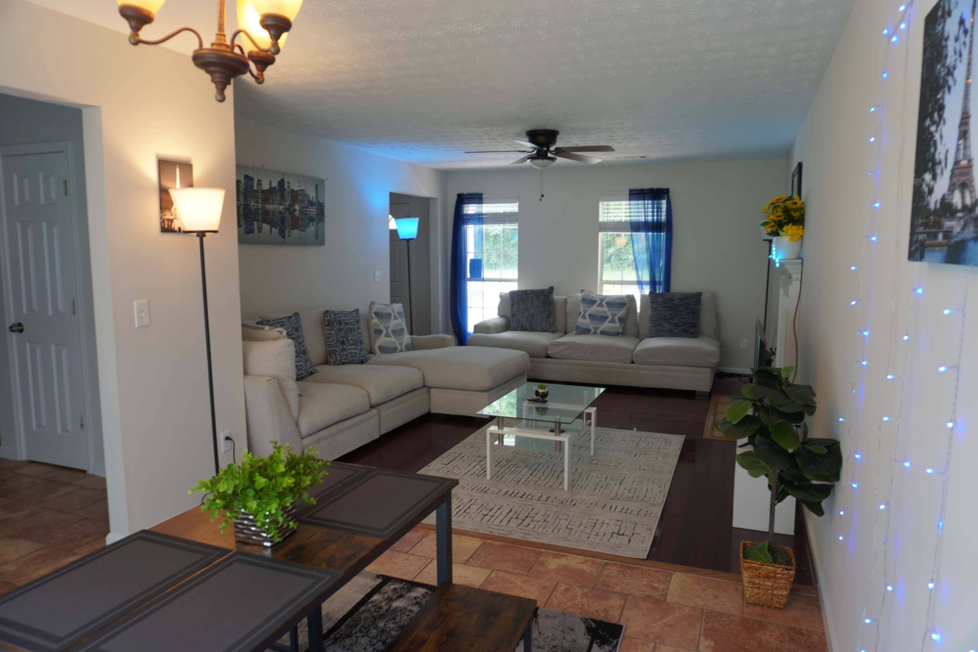 The living room features a light-colored sectional sofa, a glass coffee table, and a patterned area rug, with natural light coming through large windows.