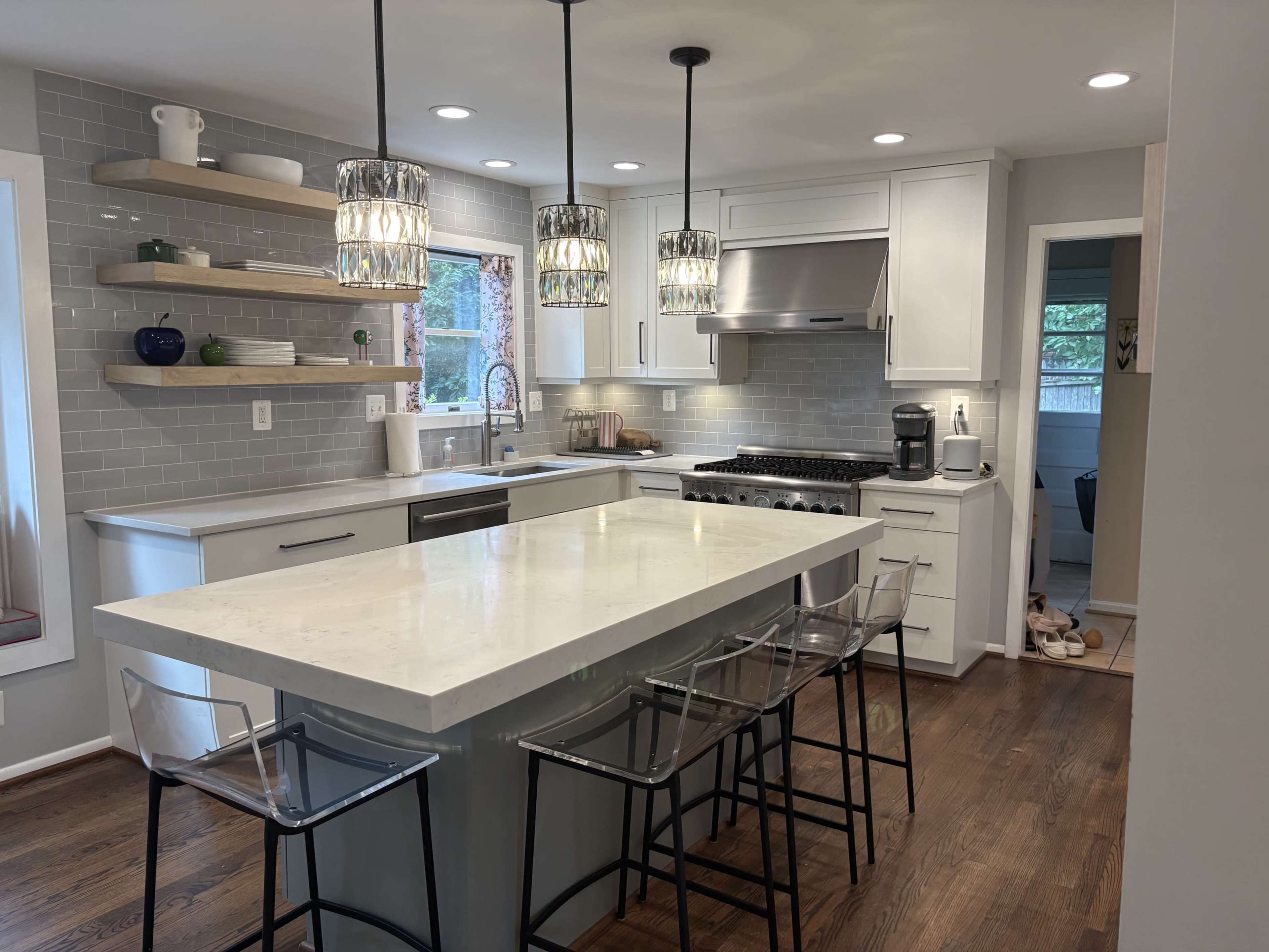 The image shows a modern kitchen with a large island featuring four transparent stools, stainless steel appliances, and overhead pendant lights.