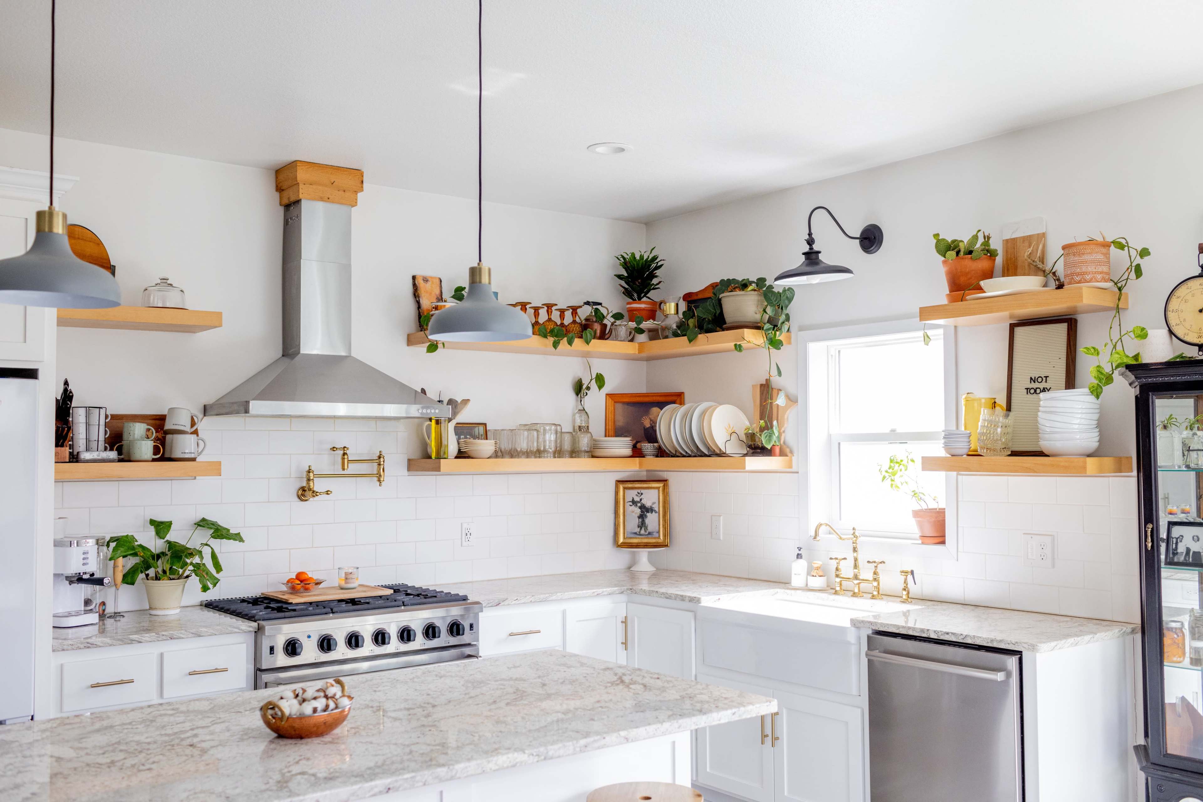 A modern kitchen features white cabinetry, open shelving with plants and dishware, and a large countertop with a stainless steel stove.