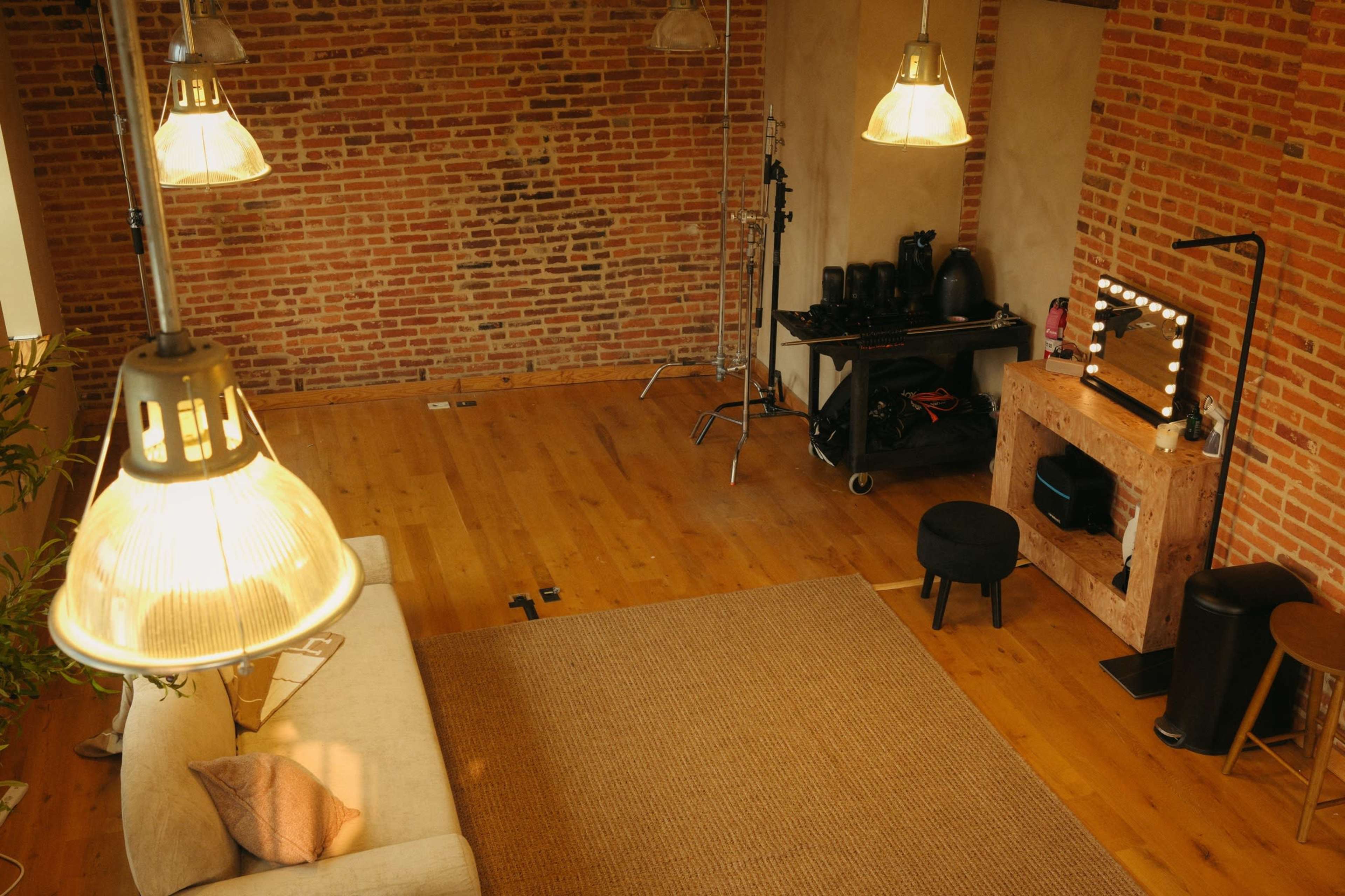 The image shows a spacious room with exposed brick walls, wooden flooring, a sofa, a rug, and a makeup station with lights.