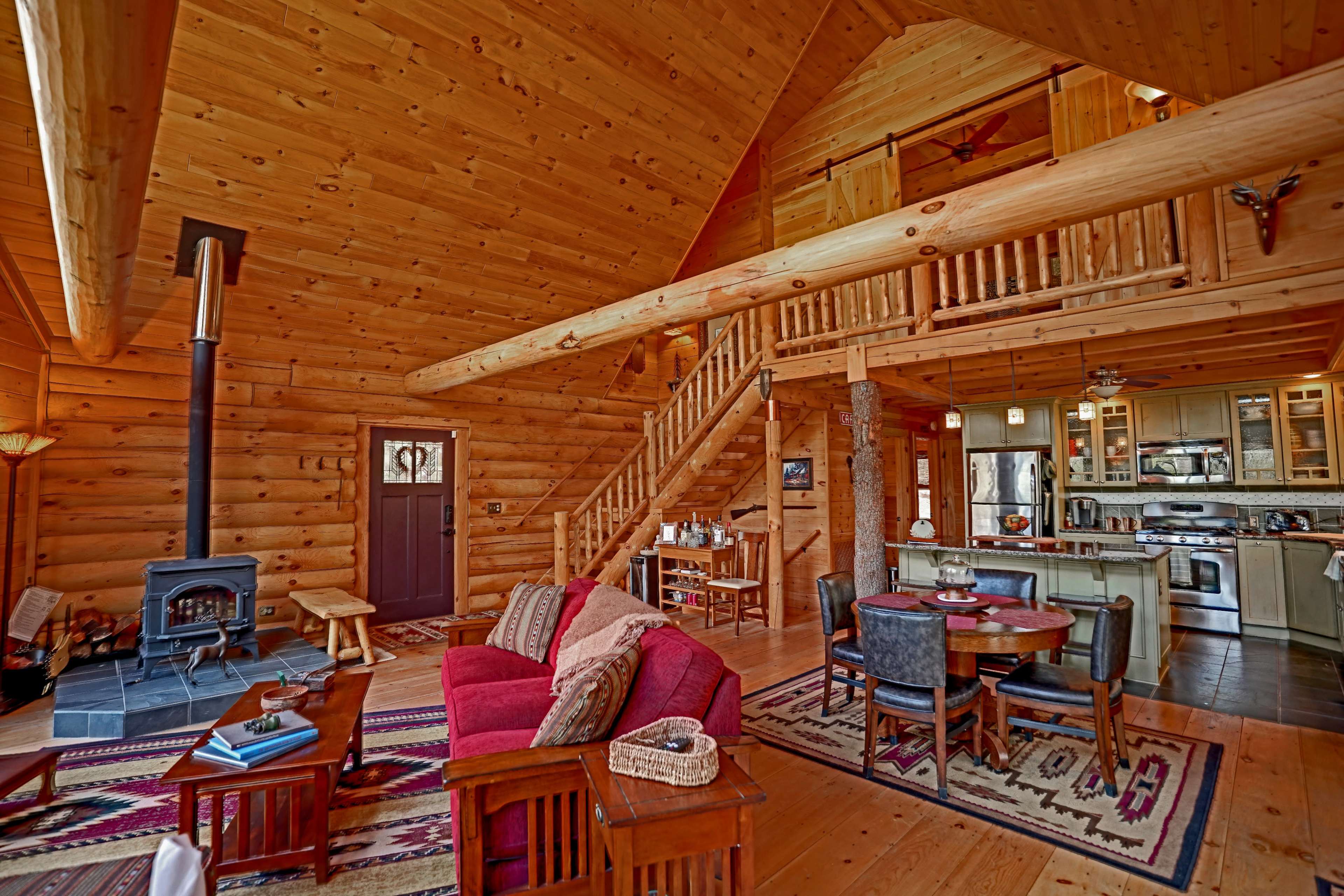 The image shows a spacious log cabin interior featuring wooden walls, a staircase, a seating area with a red couch, a dining table, and a kitchen visible in the background.