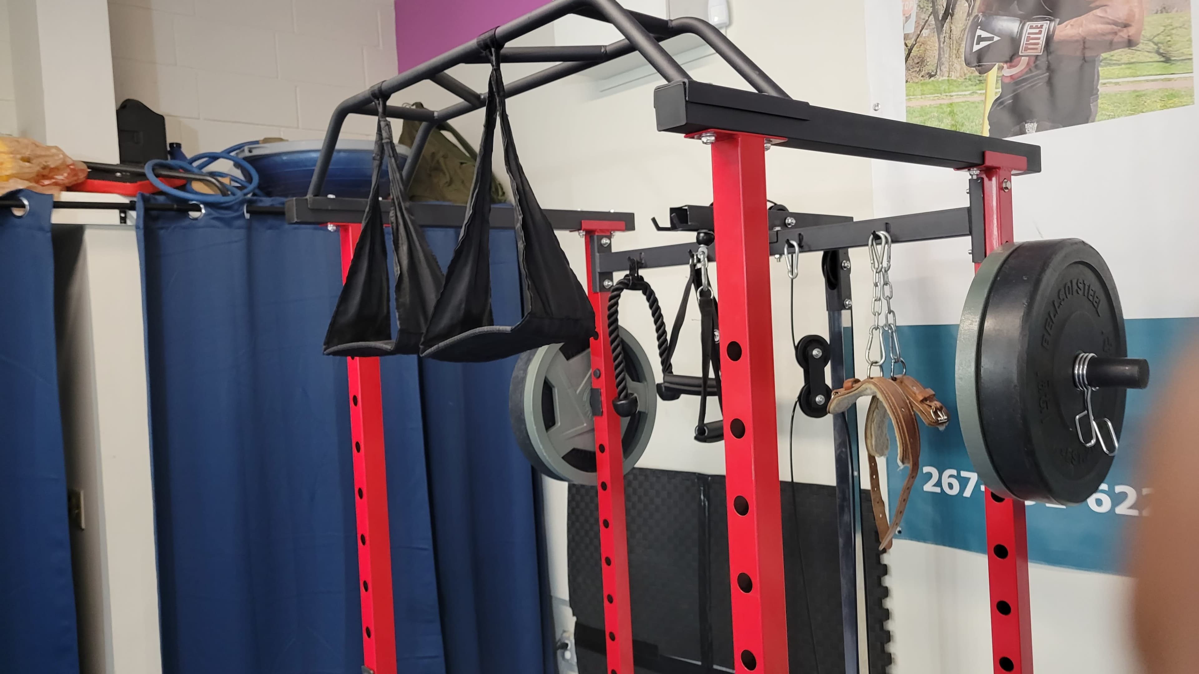 The image shows a red weightlifting rack equipped with various attachments and weights, positioned against a backdrop of blue curtains.