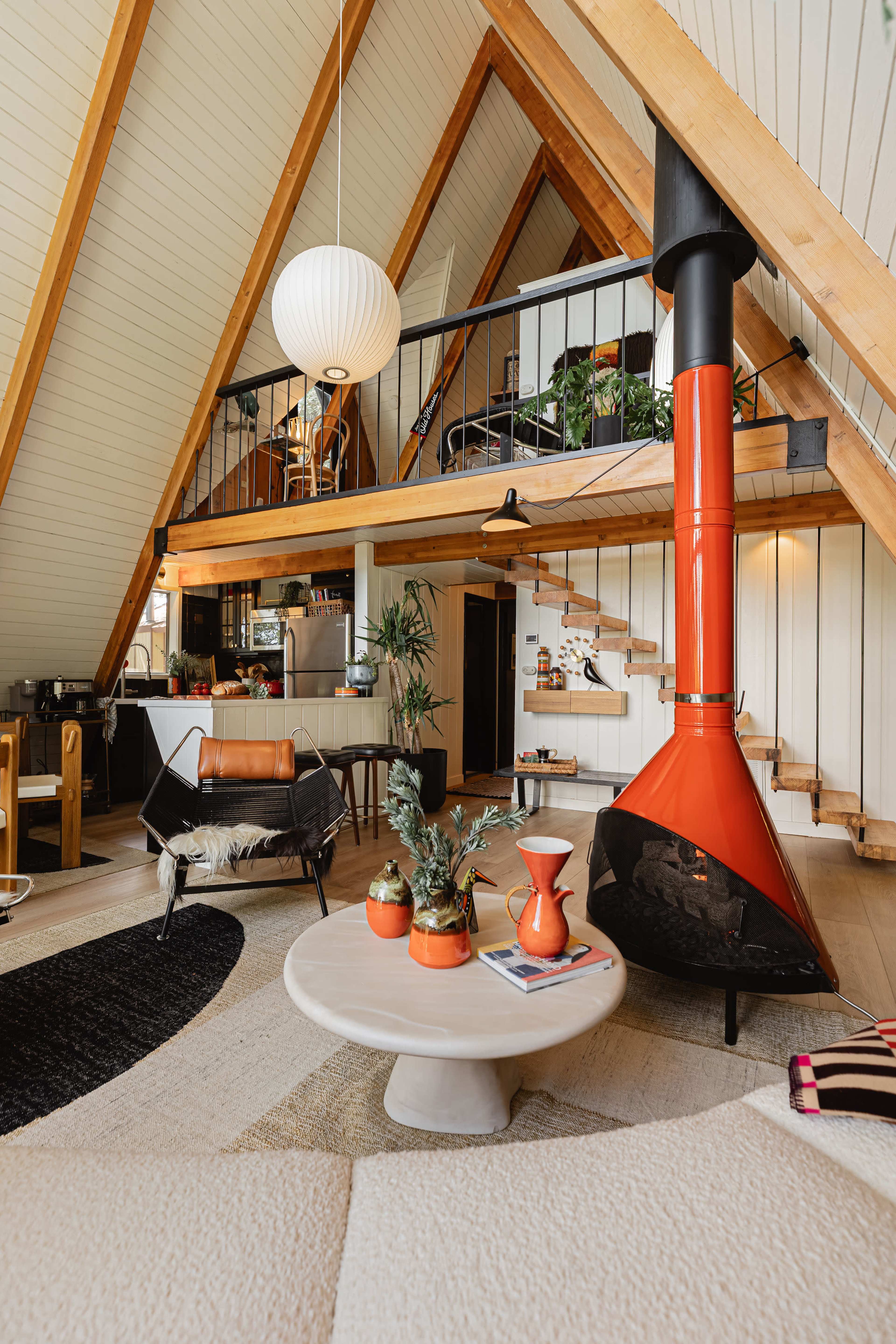The interior of a modern, A-frame house features a bright, open living space with wooden beams, an orange fireplace, and a mix of furniture and decor elements.