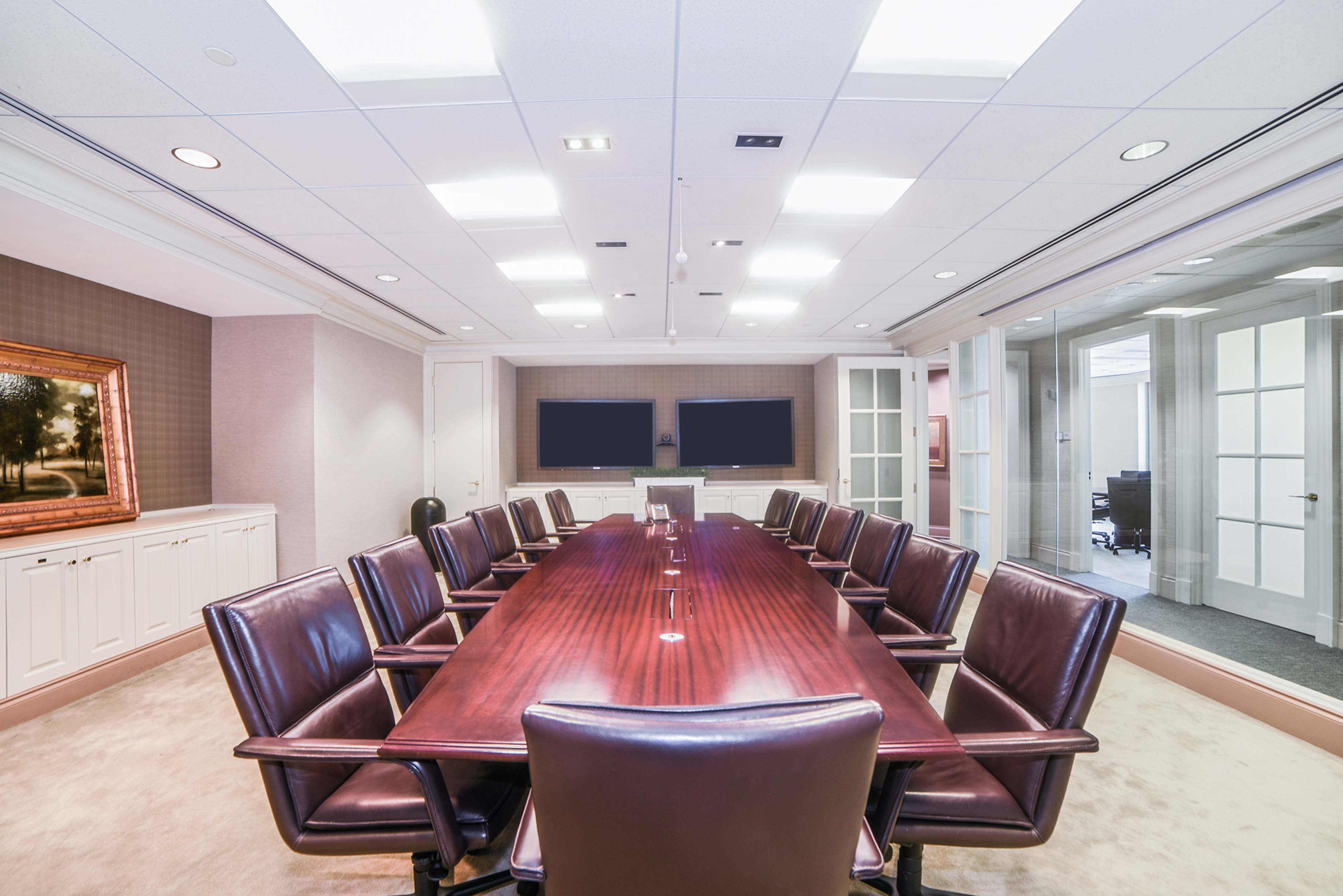 The image shows a conference room with a long wooden table surrounded by leather chairs, two large screens mounted on the wall, and glass doors leading to an adjacent office space.