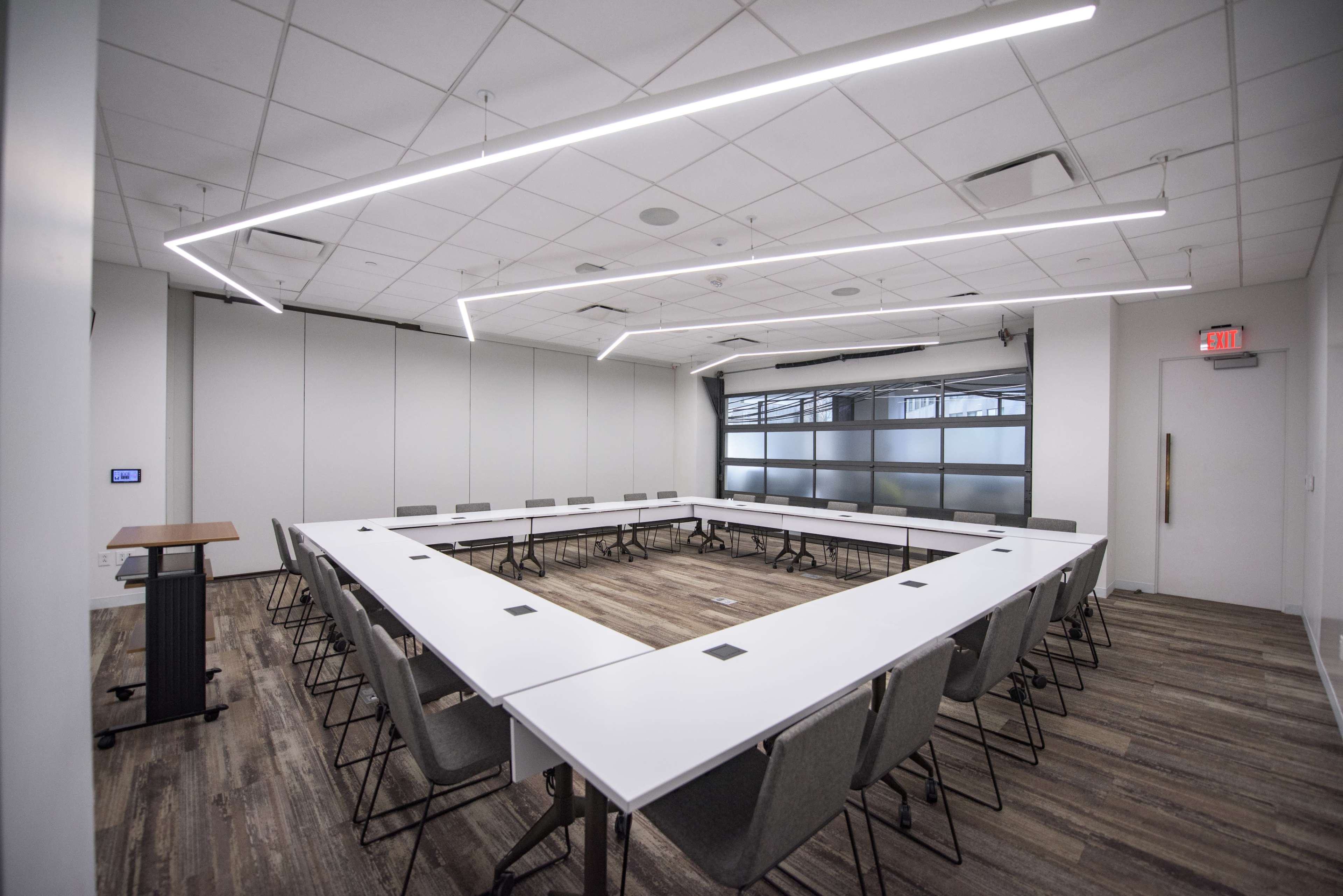 A conference room features a large, rectangular table surrounded by chairs, with modern lighting and large windows.