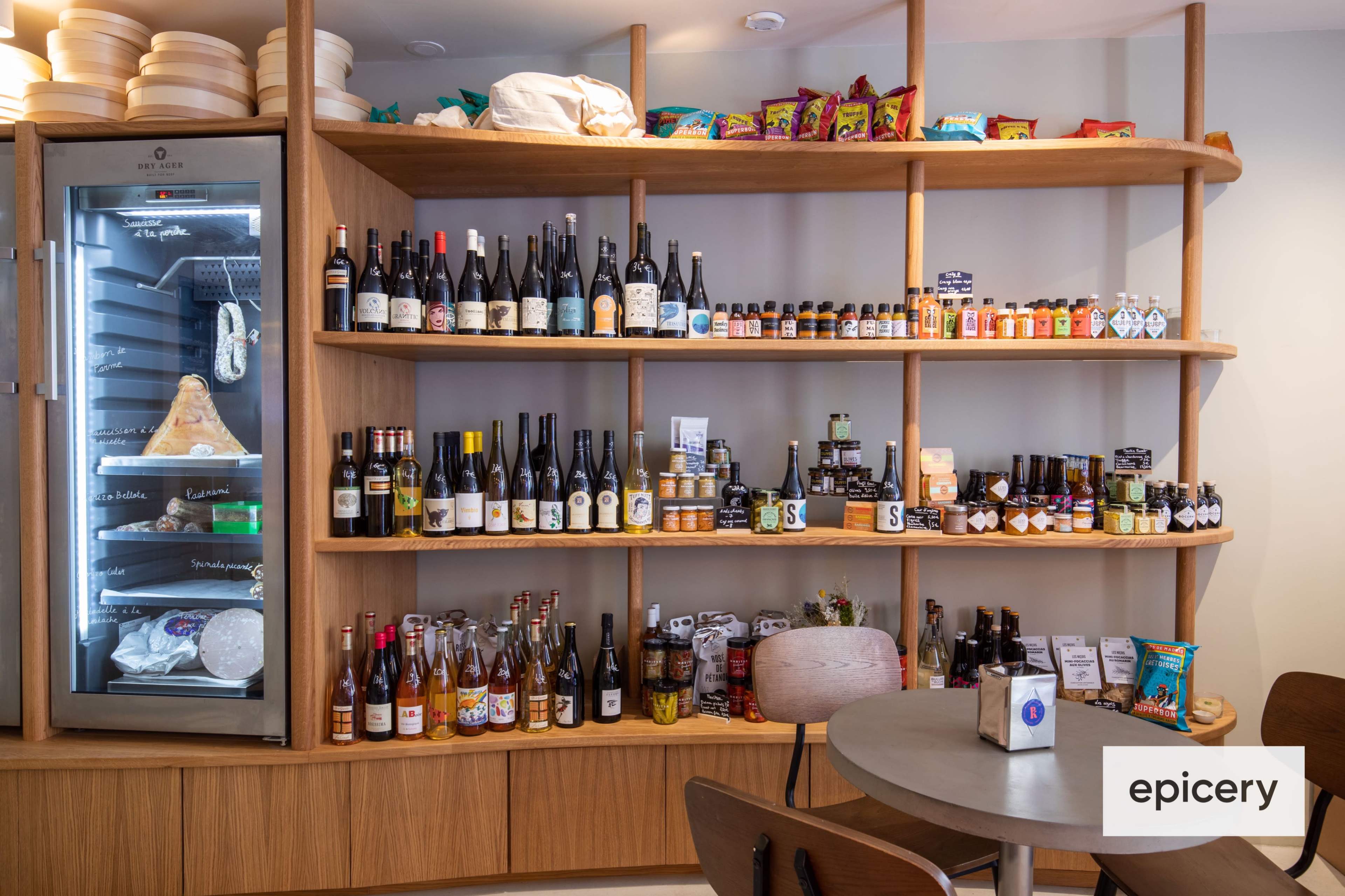 A wooden shelf displays various bottles, jars, and packaged goods, with a glass-front refrigerator beside it in a food retail space.