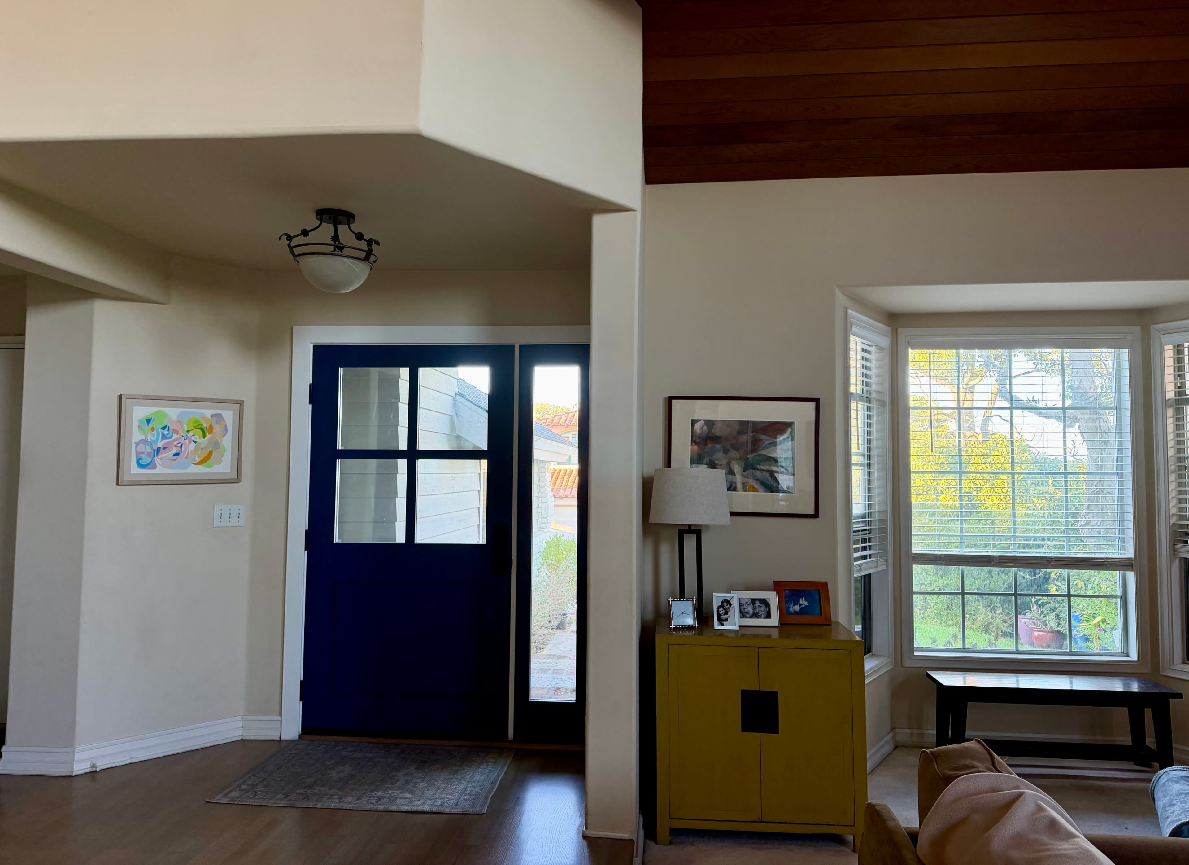 The image shows a well-lit entryway featuring a blue front door, a side window, and a yellow cabinet with framed pictures.