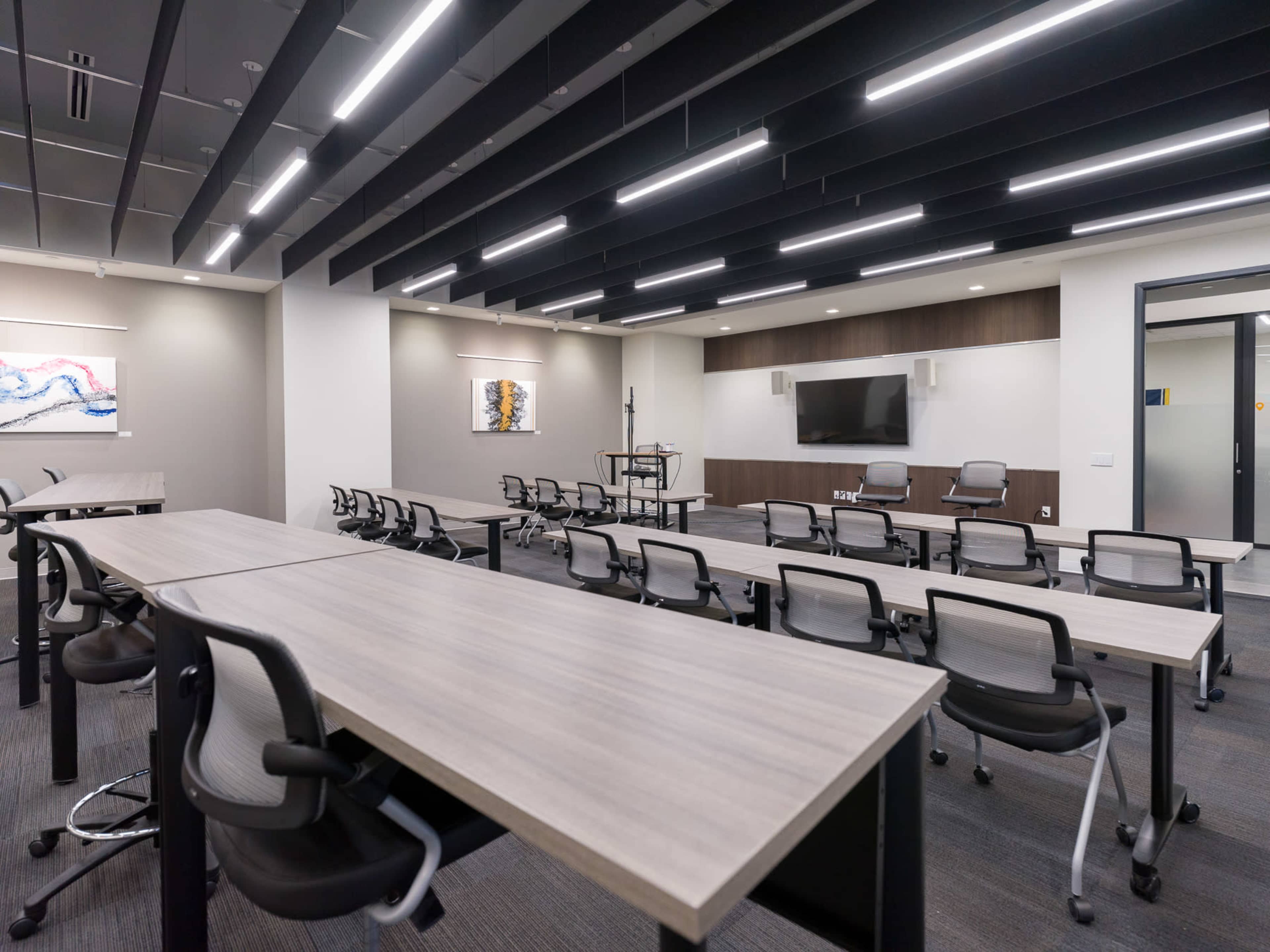 The image shows a modern conference room with arranged tables and chairs, featuring a wall-mounted television and artwork.
