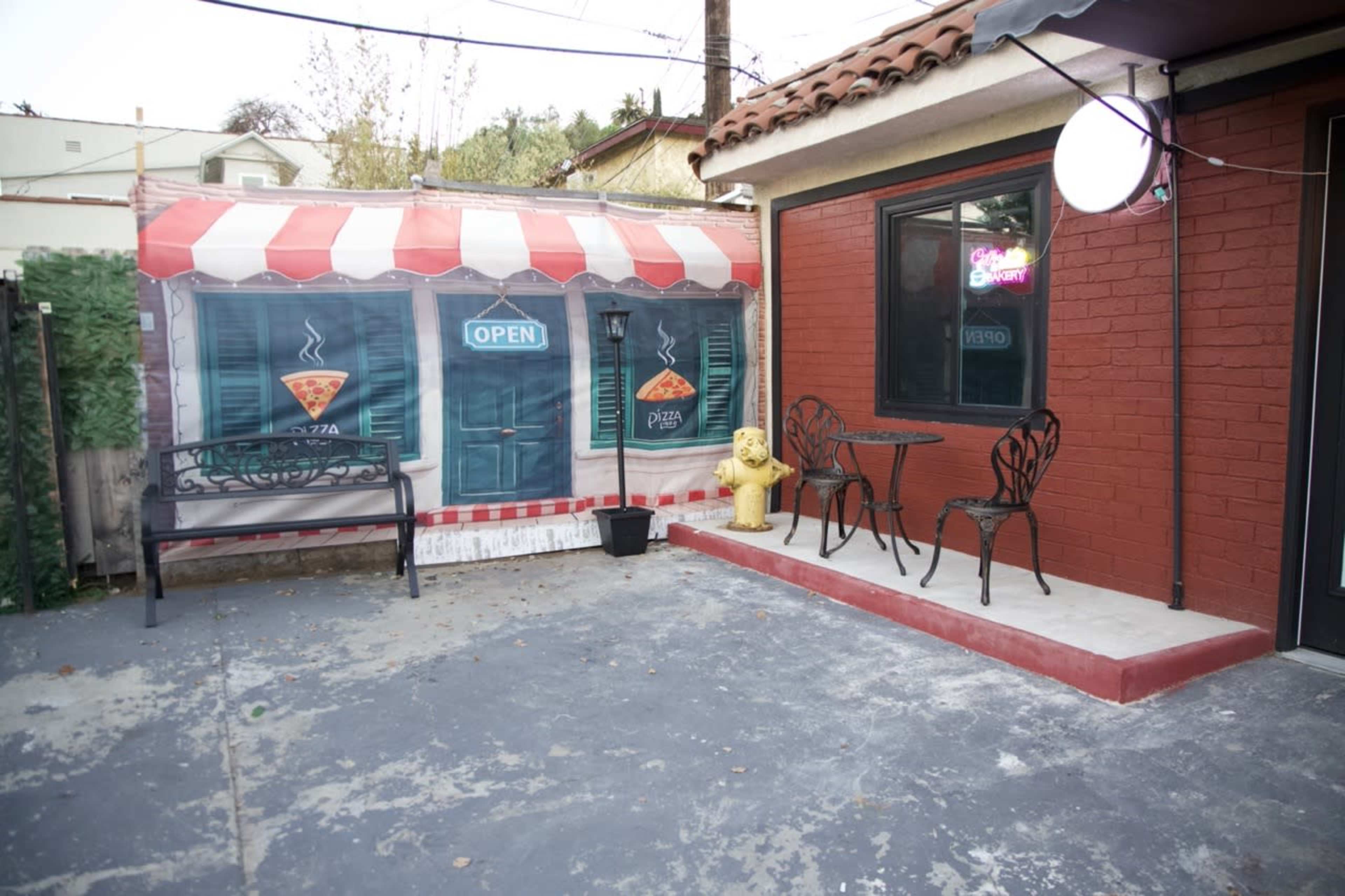 The image shows an outdoor dining area with a small table and two chairs next to a graphic storefront featuring pizza signage.