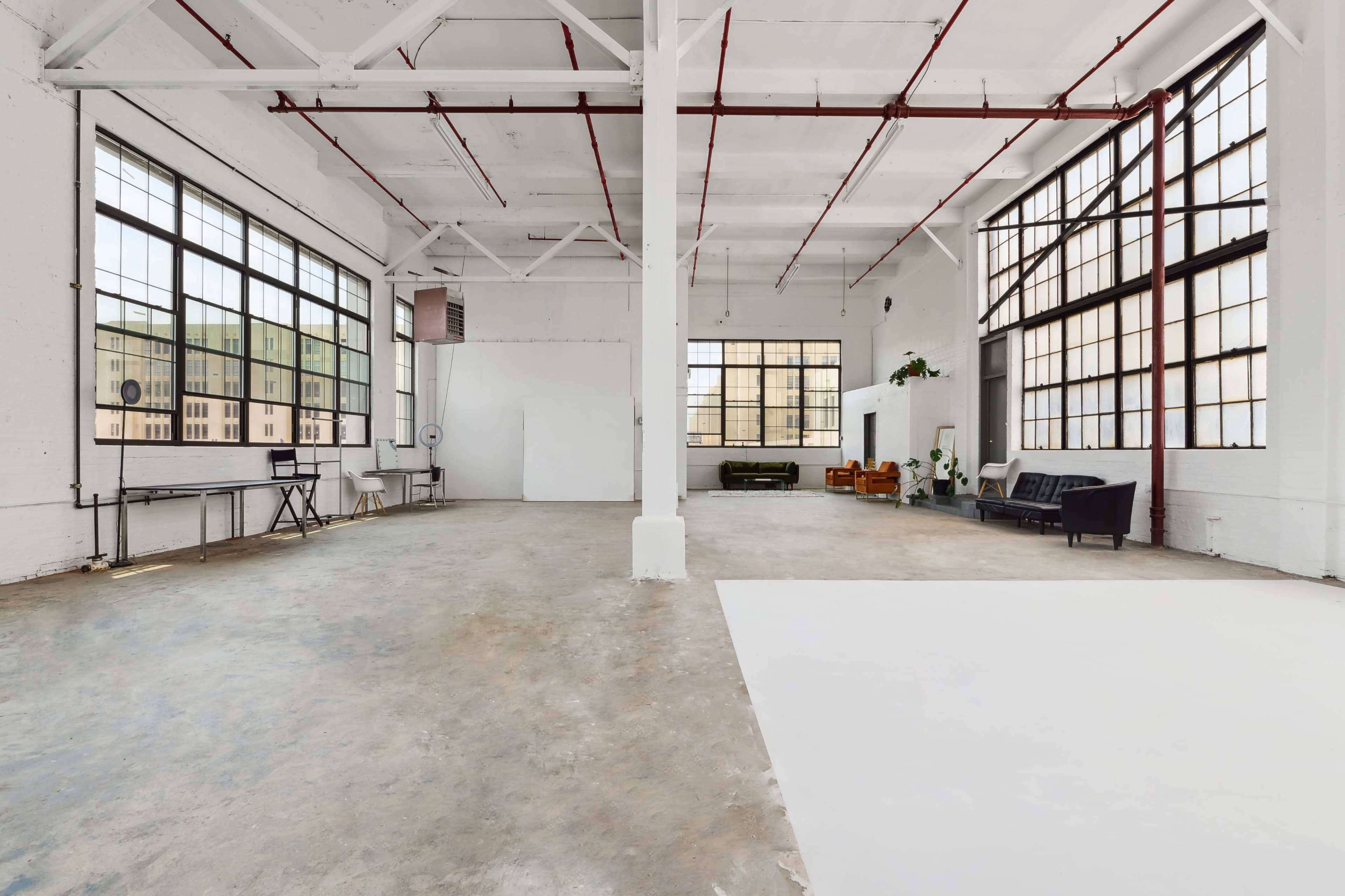 A spacious, empty industrial-style room with large windows and minimal furnishings, featuring concrete floors and white walls.