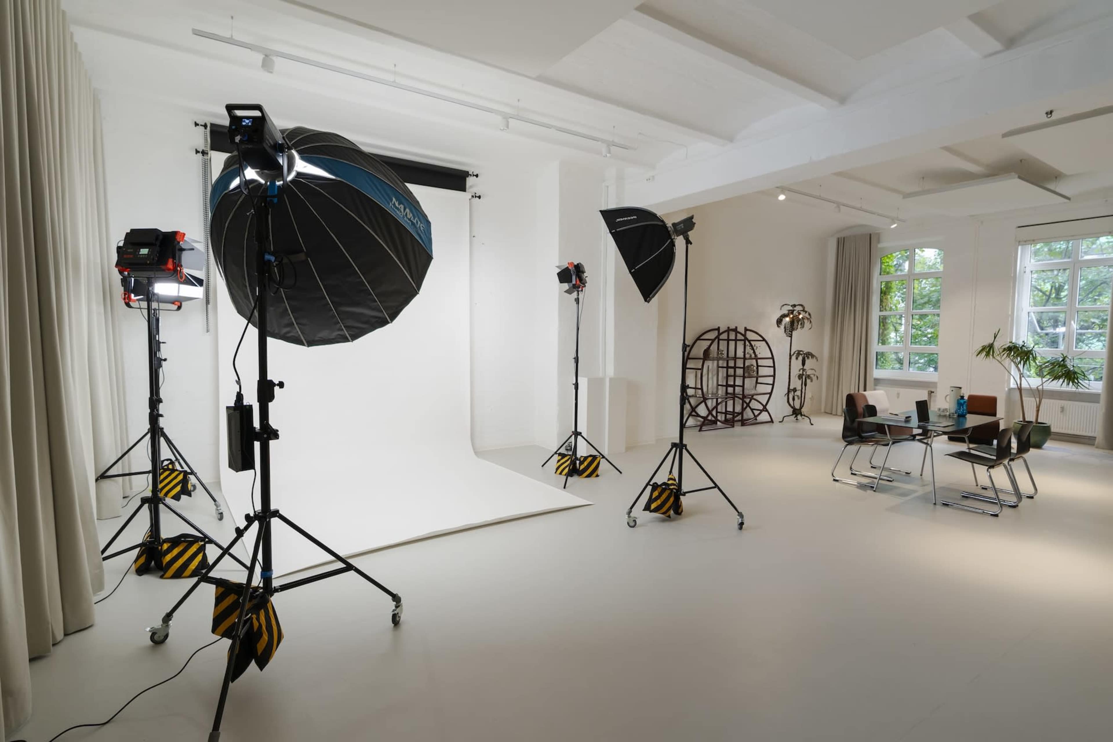 The image shows a well-lit studio setup featuring multiple softbox lights and a backdrop in an open space with furniture and plants.