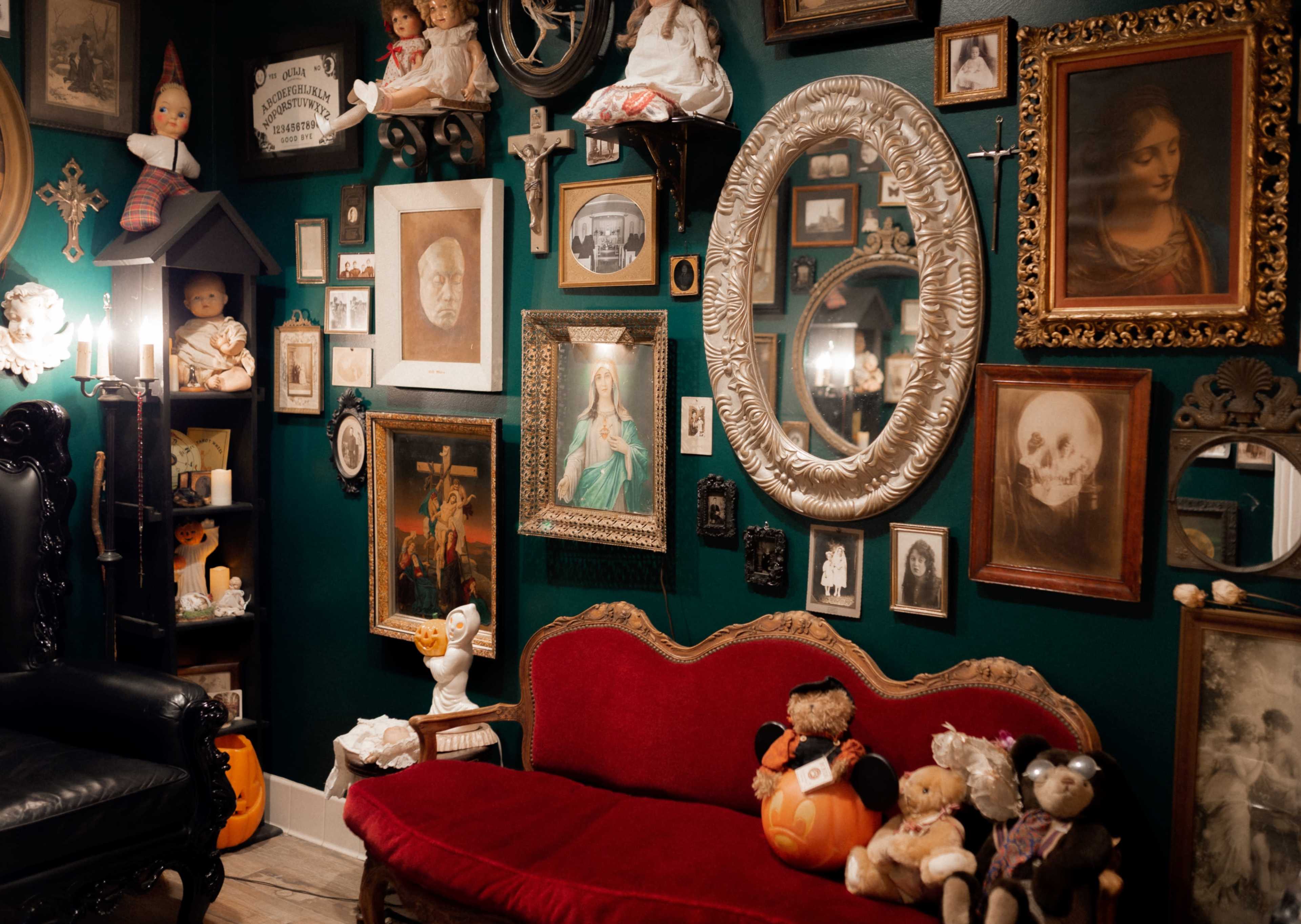 The image depicts a cozy room with a red upholstered couch surrounded by an eclectic collection of framed artwork, dolls, and decorative items on the walls.