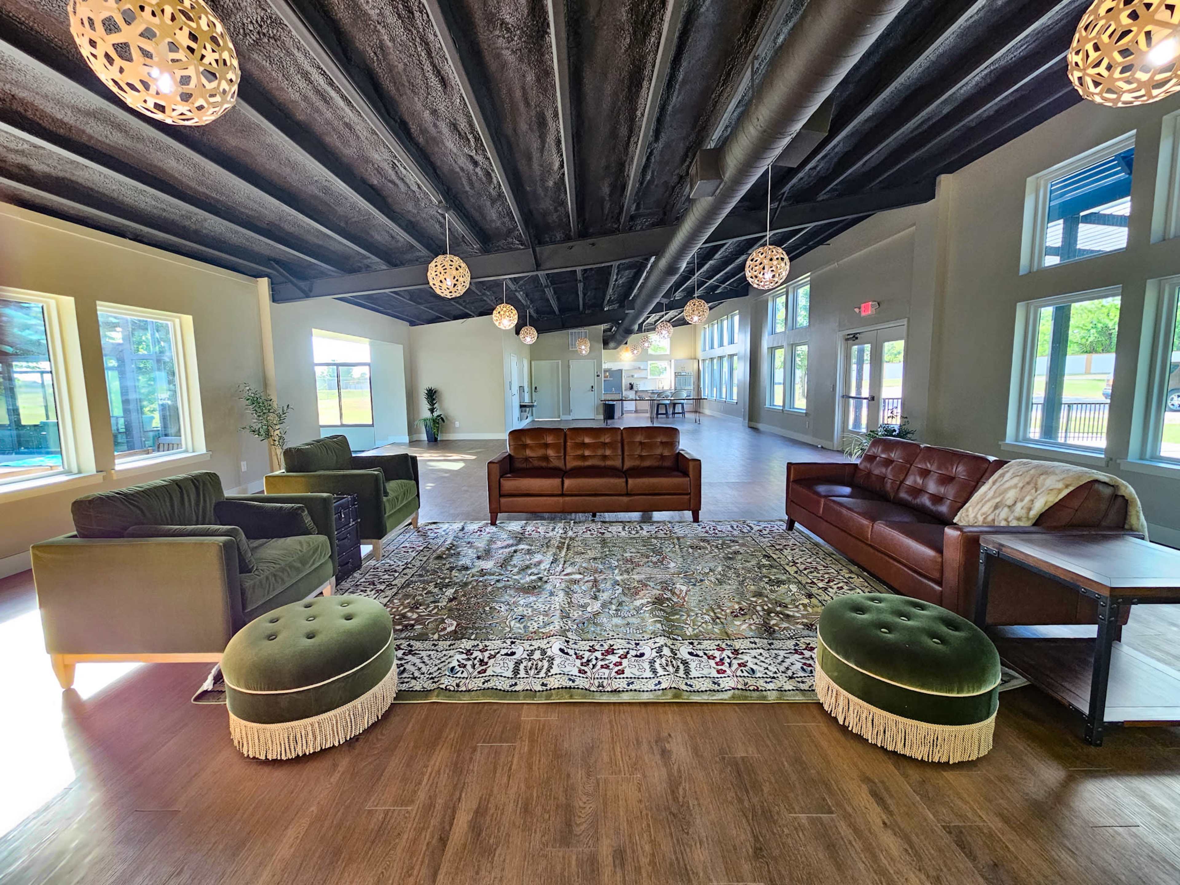 The image shows a spacious lounge area featuring multiple sofas, decorative poufs, and large windows that allow natural light to fill the room.