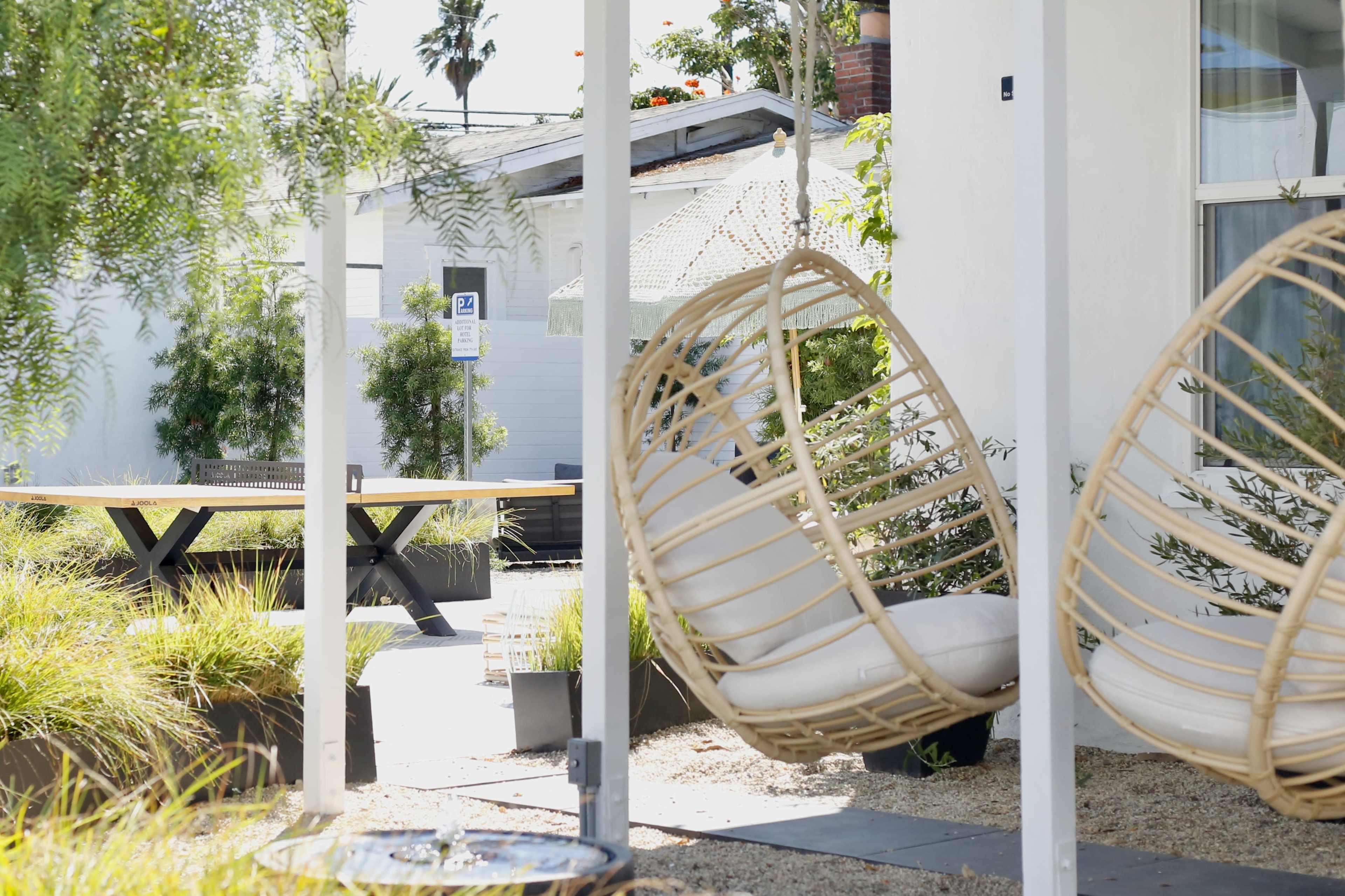 The image shows a tranquil outdoor seating area featuring two hanging chairs, a wooden table in the background, and landscaped greenery.