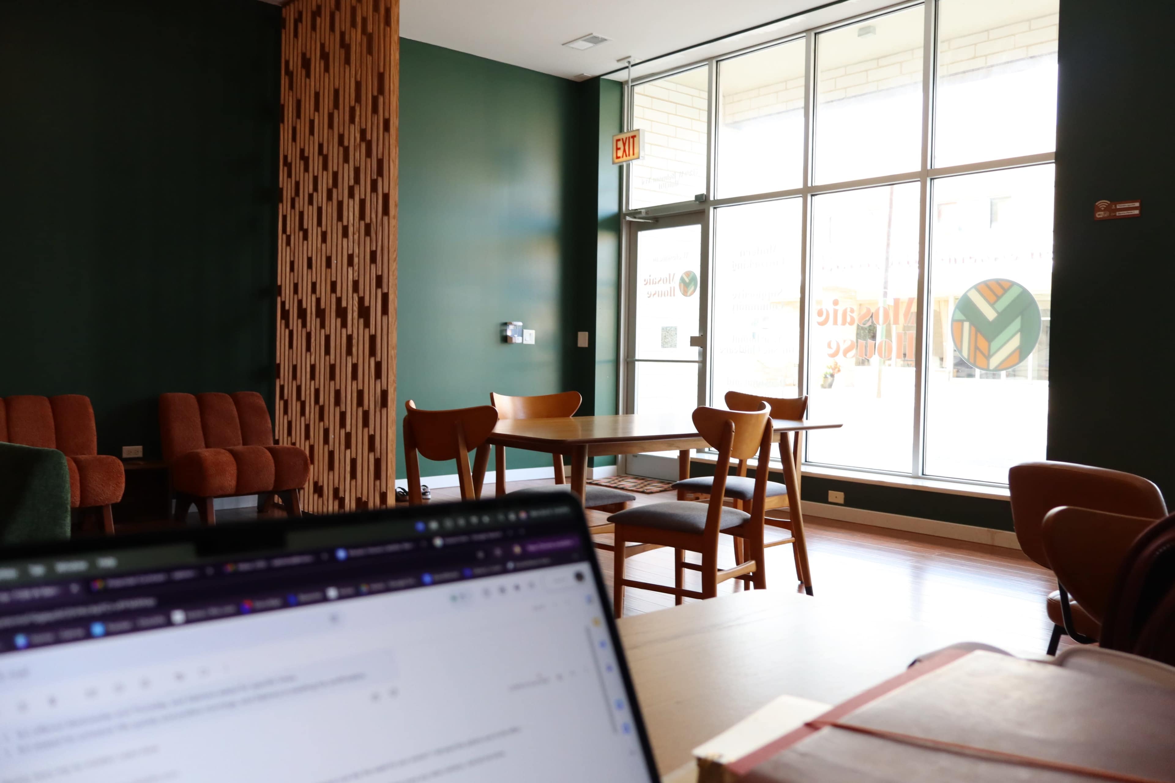 The image shows an indoor café or workspace with a green wall, wooden furniture, and large windows letting in natural light.