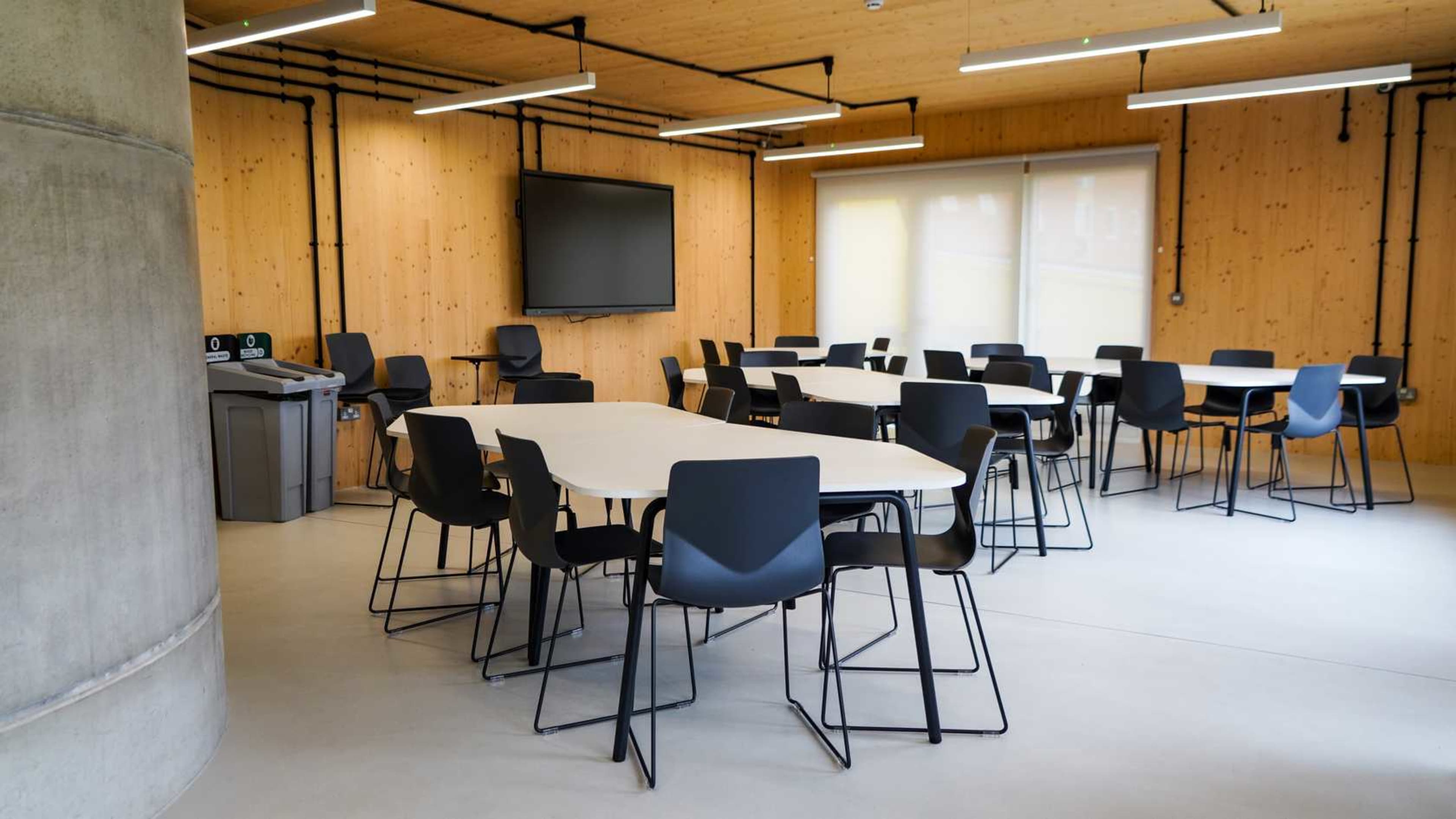 The image shows a modern classroom with several round tables and black chairs, a projector screen on the wall, and large windows with blinds.