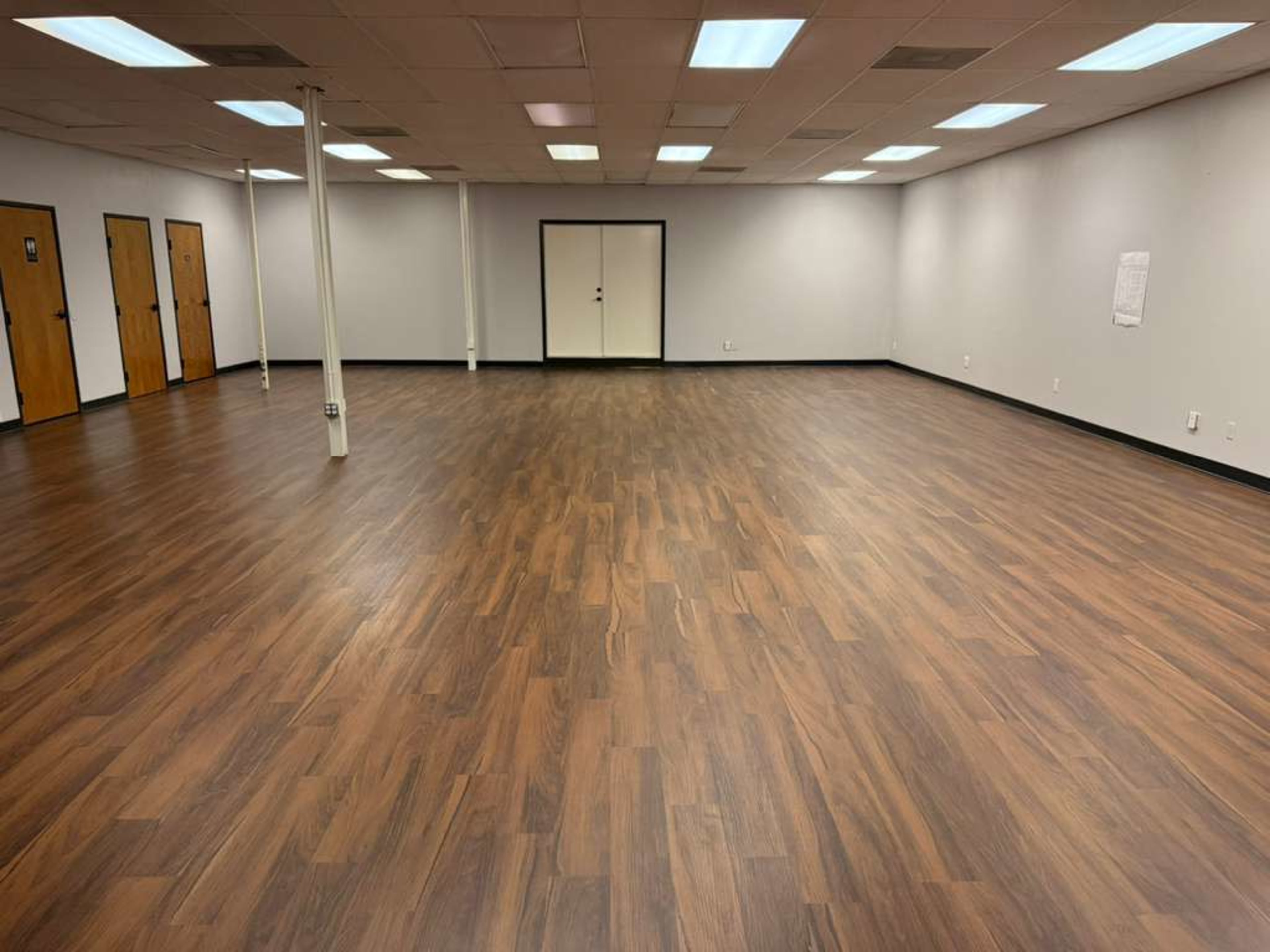 The image shows a spacious, empty room with wooden flooring, several doors along the walls, and overhead fluorescent lights.