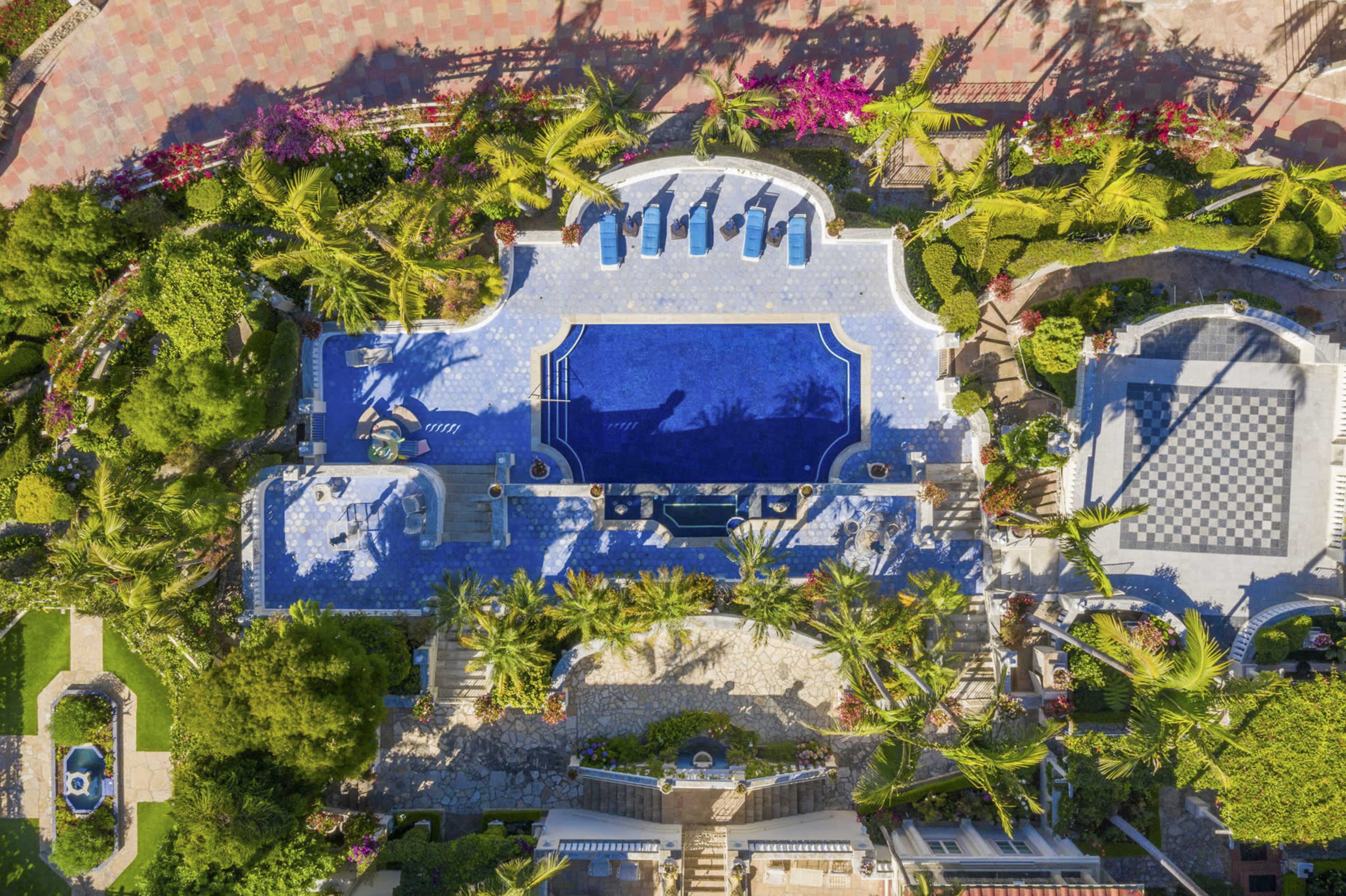 The image shows an aerial view of a luxury villa with a large blue swimming pool, surrounded by palm trees and landscaped gardens.