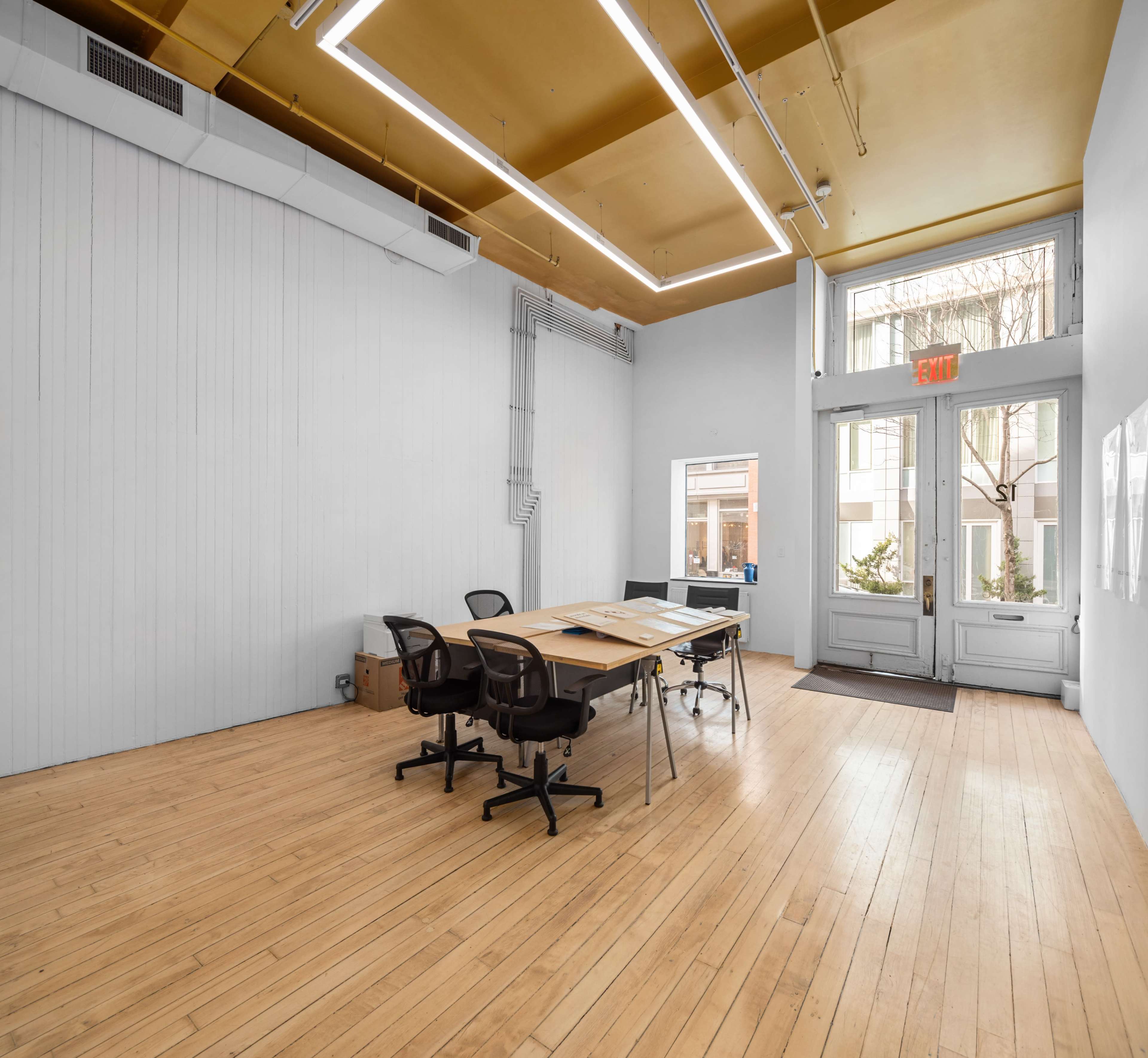A spacious, empty room with wooden flooring, a large table and chairs arranged for a meeting, and large windows at the entrance.