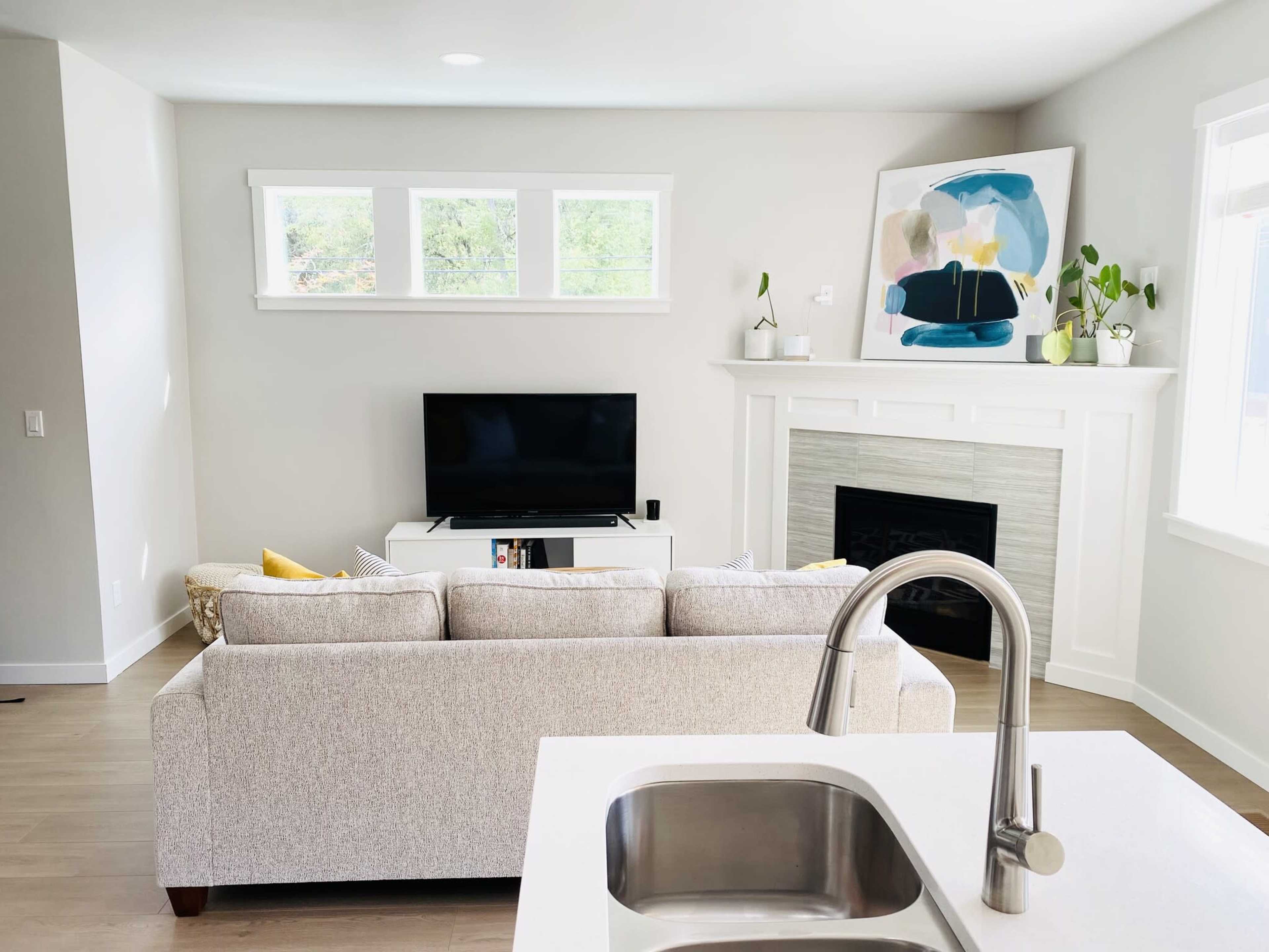 A light-filled living room features a beige sofa facing a television on a white media console, with a modern fireplace and artwork on the wall.
