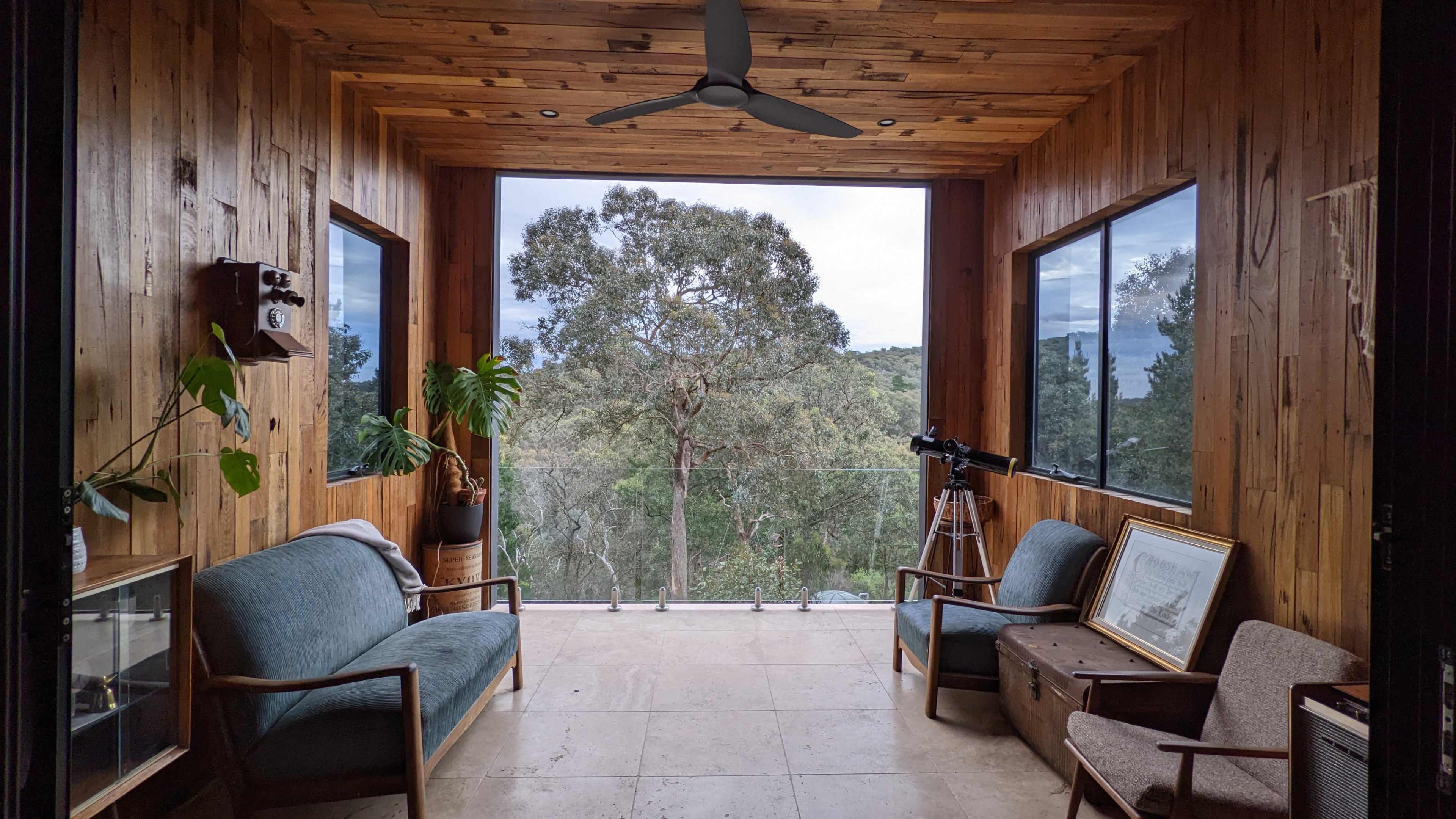 The image depicts a wooden-paneled room with large windows overlooking a natural landscape, featuring two couches, a telescope, and a few pieces of furniture.