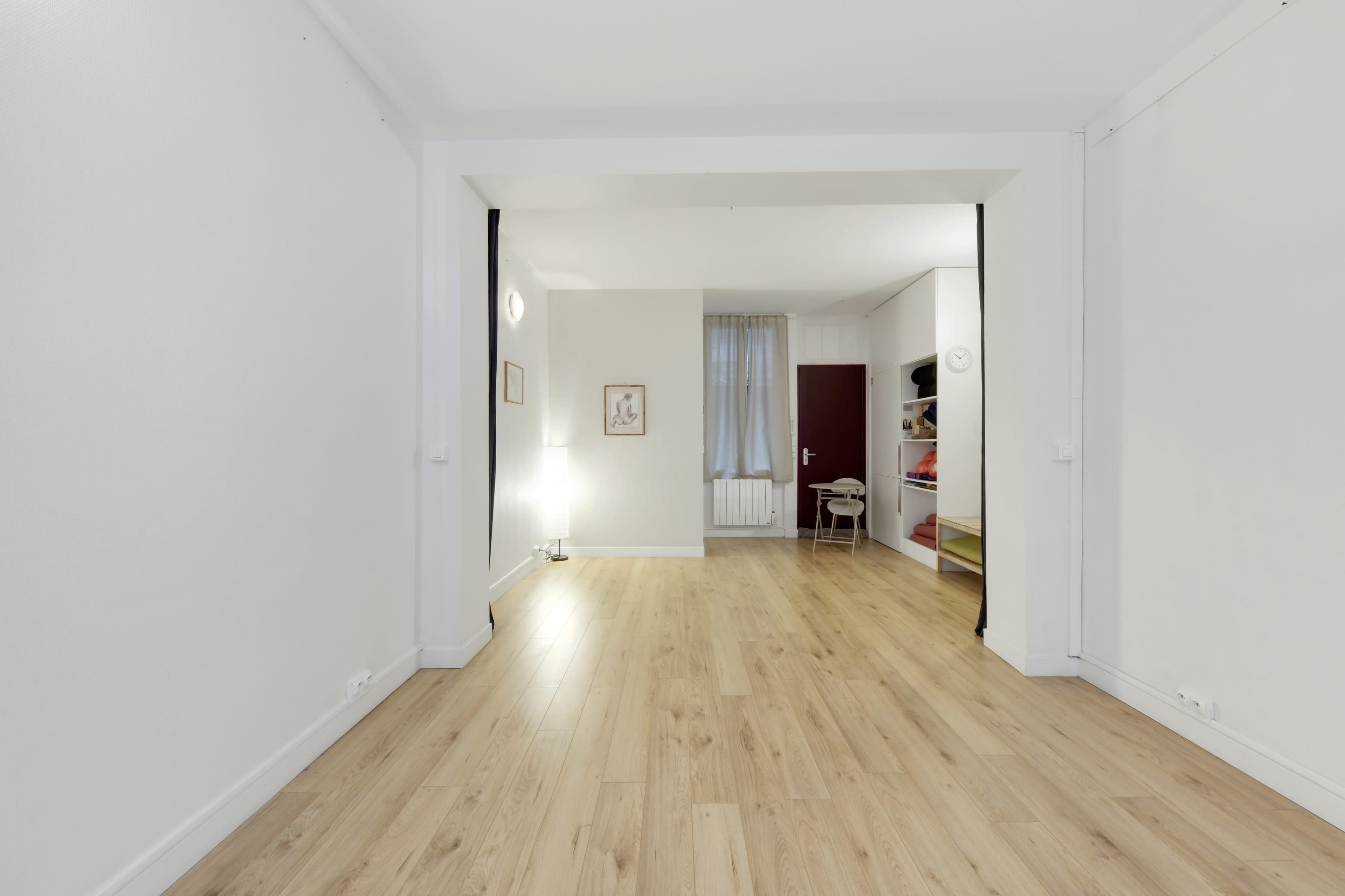 The image shows a spacious, empty room with light wooden flooring, a lamp in the corner, and an open doorway leading to a well-lit area with a small chair and shelves.