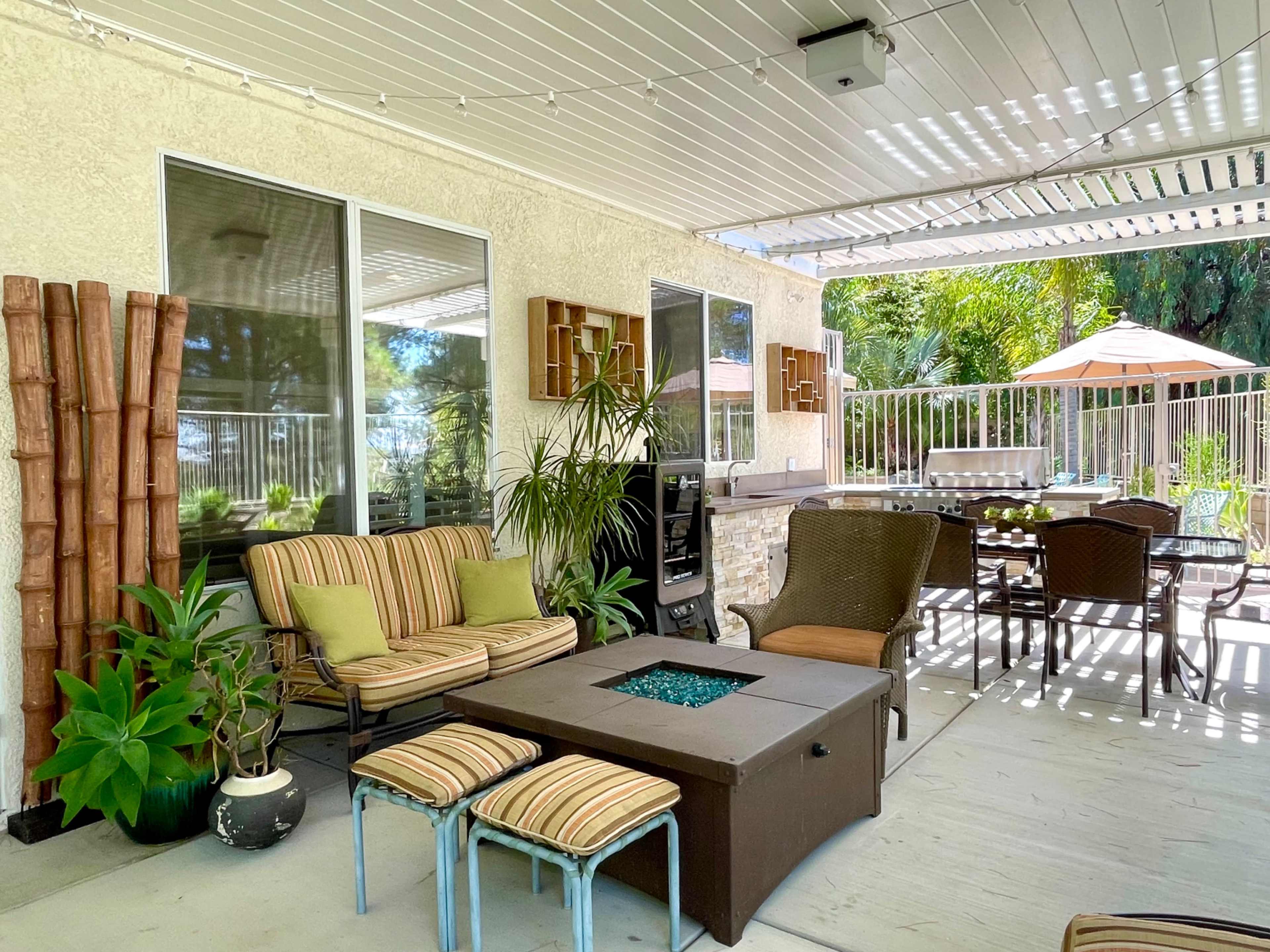 A shaded outdoor patio with seating, a fire table, and a dining area surrounded by plants and patio furniture.