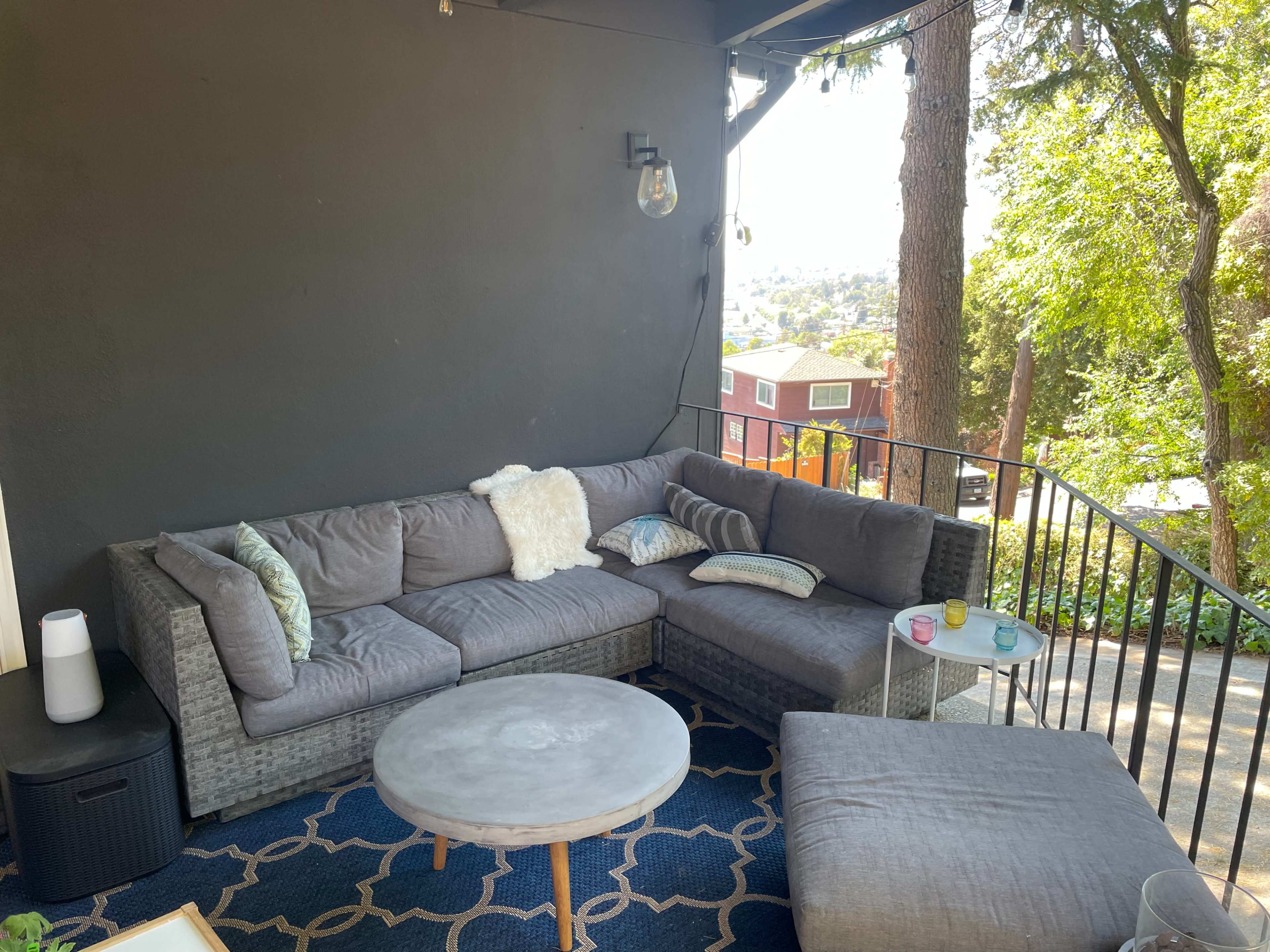 A cozy outdoor seating area with a gray sectional sofa, a round coffee table, and greenery in the background.