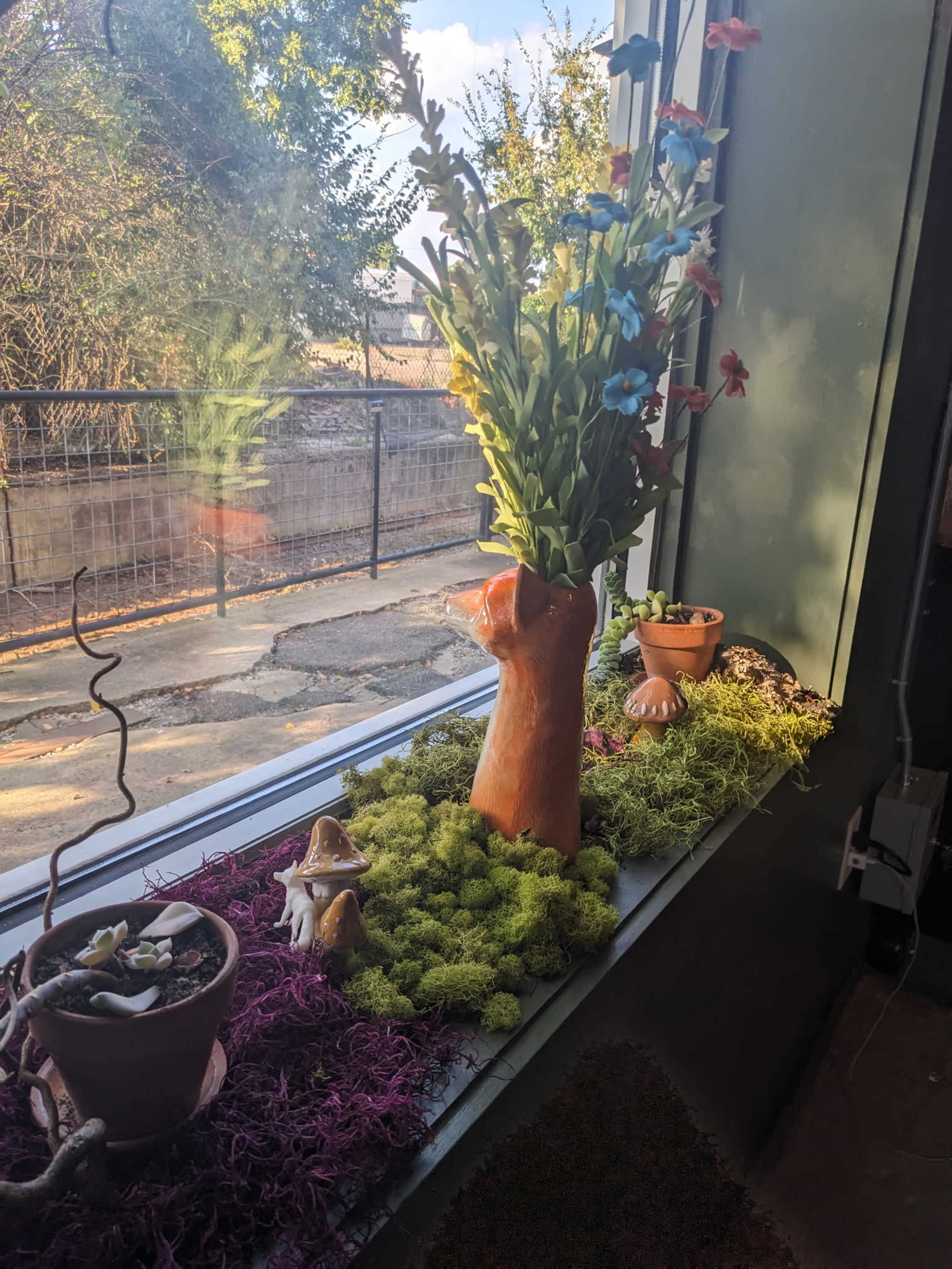A windowsill displays a vibrant arrangement of faux flowers in a decorative vase alongside small potted plants and decorative figurines set on a bed of green moss.
