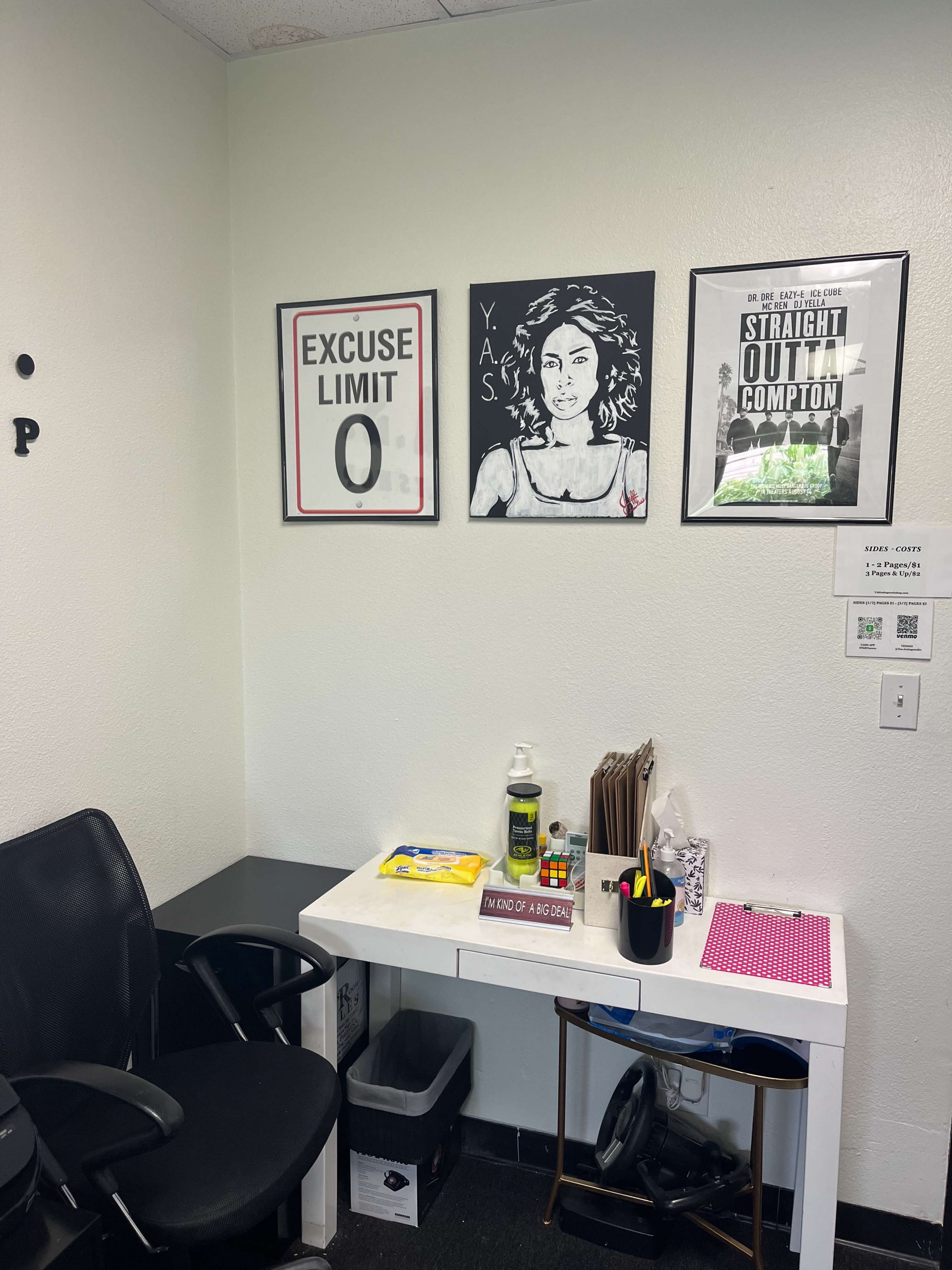 A small office space featuring a desk with papers, a chair, and framed artworks on the wall.